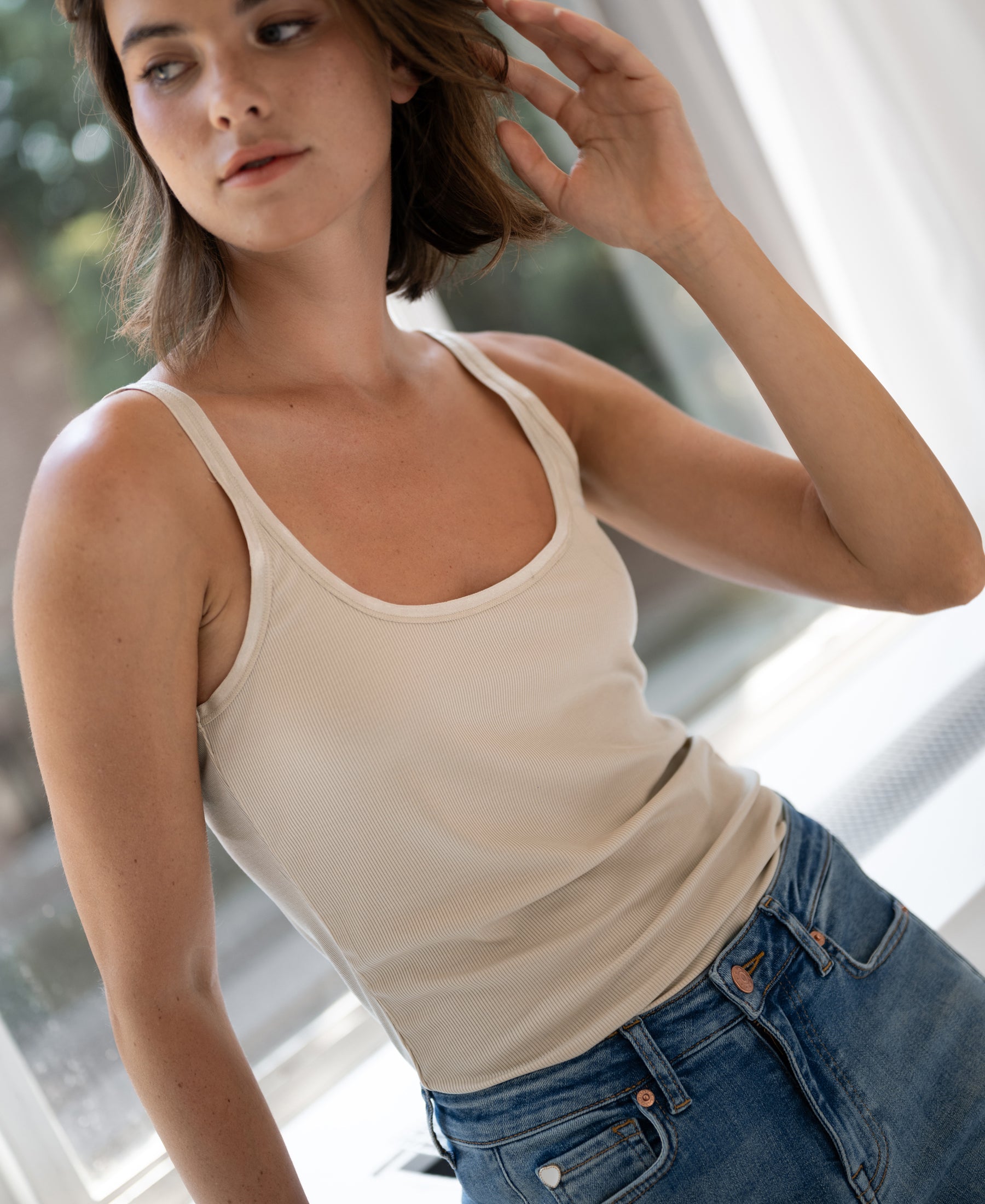 Woman wearing a Luxe Cami top in sand color stands by a bright window, touching her hair.