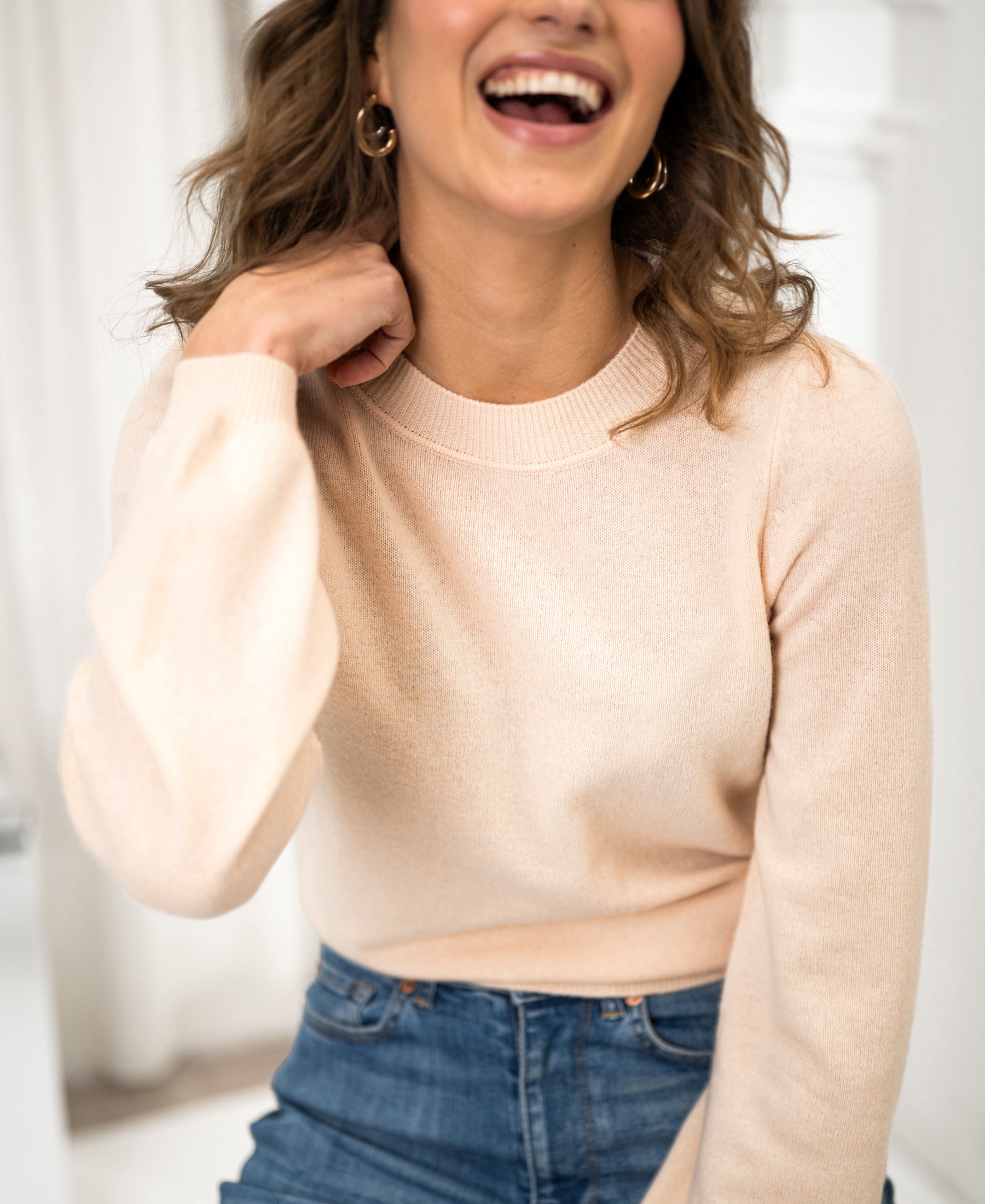 Woman wearing a PLEIN PUBLIQUE LA BAYONNE merino wool sweater in butter color, smiling indoors with hand near her neck.