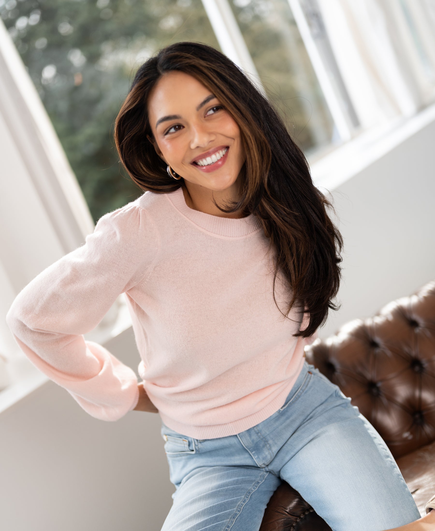 Pink merino wool sweater LA BAYONNE by PLEIN PUBLIQUE, shown on a woman with long dark hair, seated on a brown sofa.