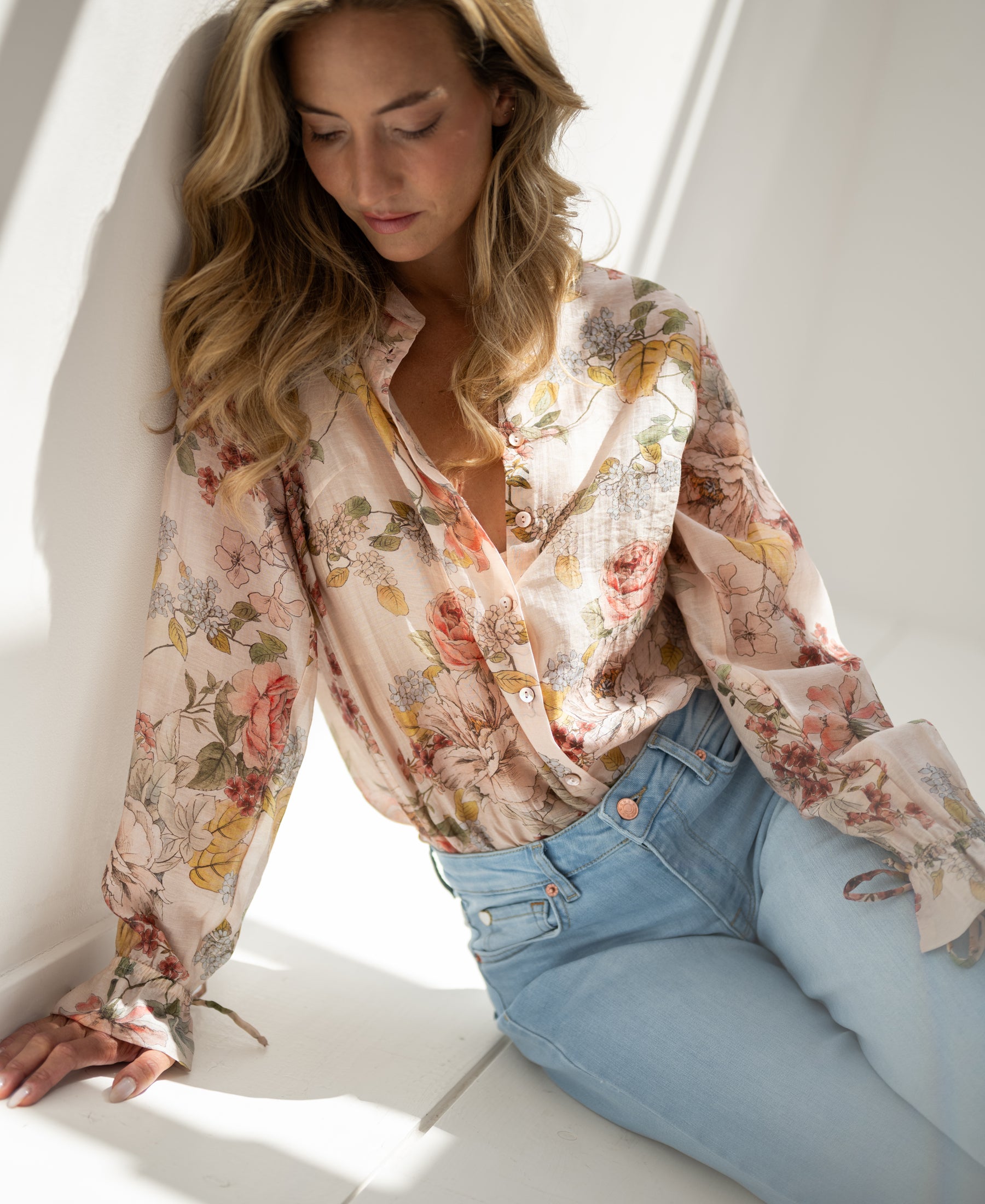 Blush floral print blouse with botanical pattern, shown worn by a woman seated near a sunlit wall.