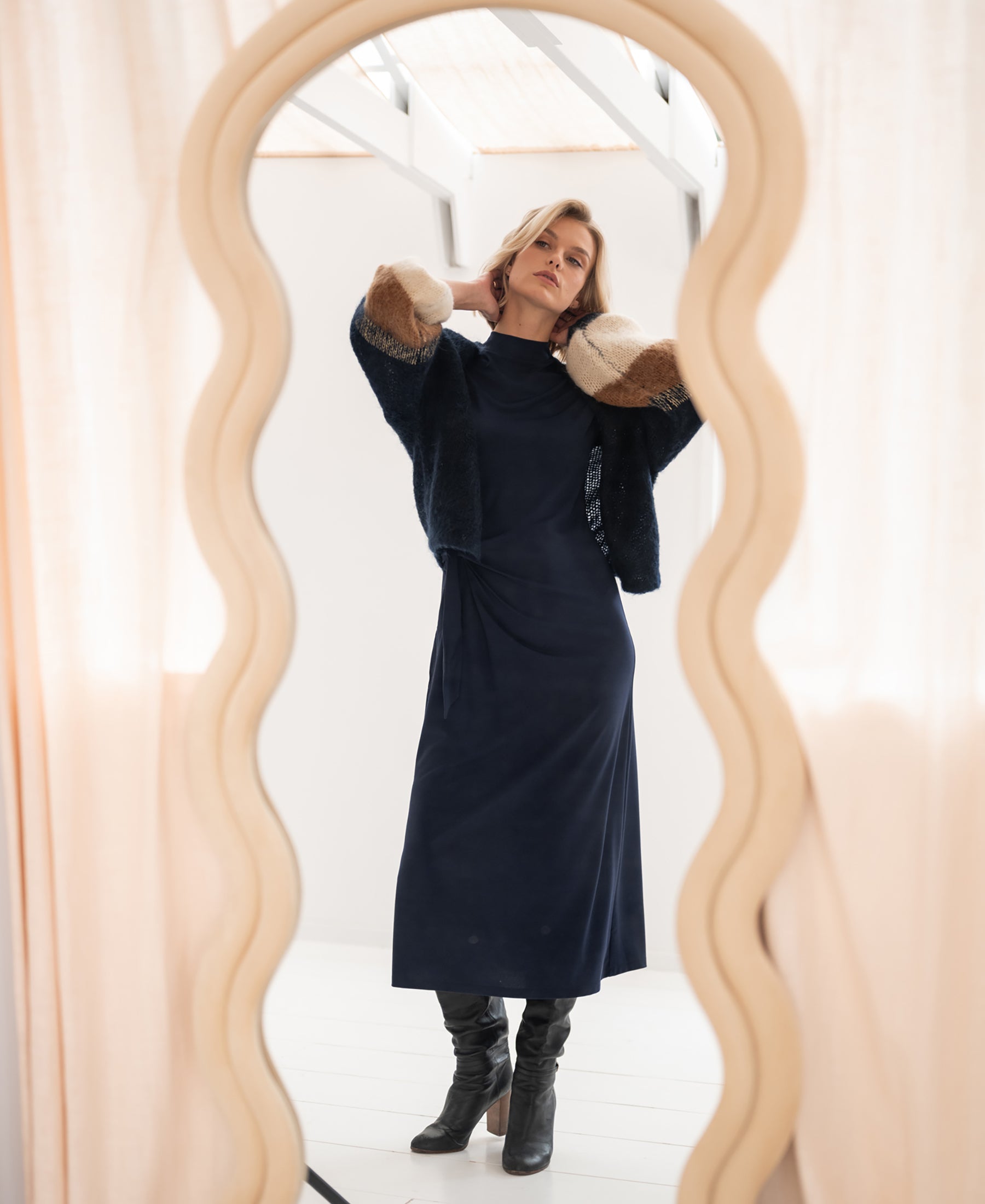 Woman wearing a PLEIN PUBLIQUE dark blue travel dress stands by a cream wavy mirror.