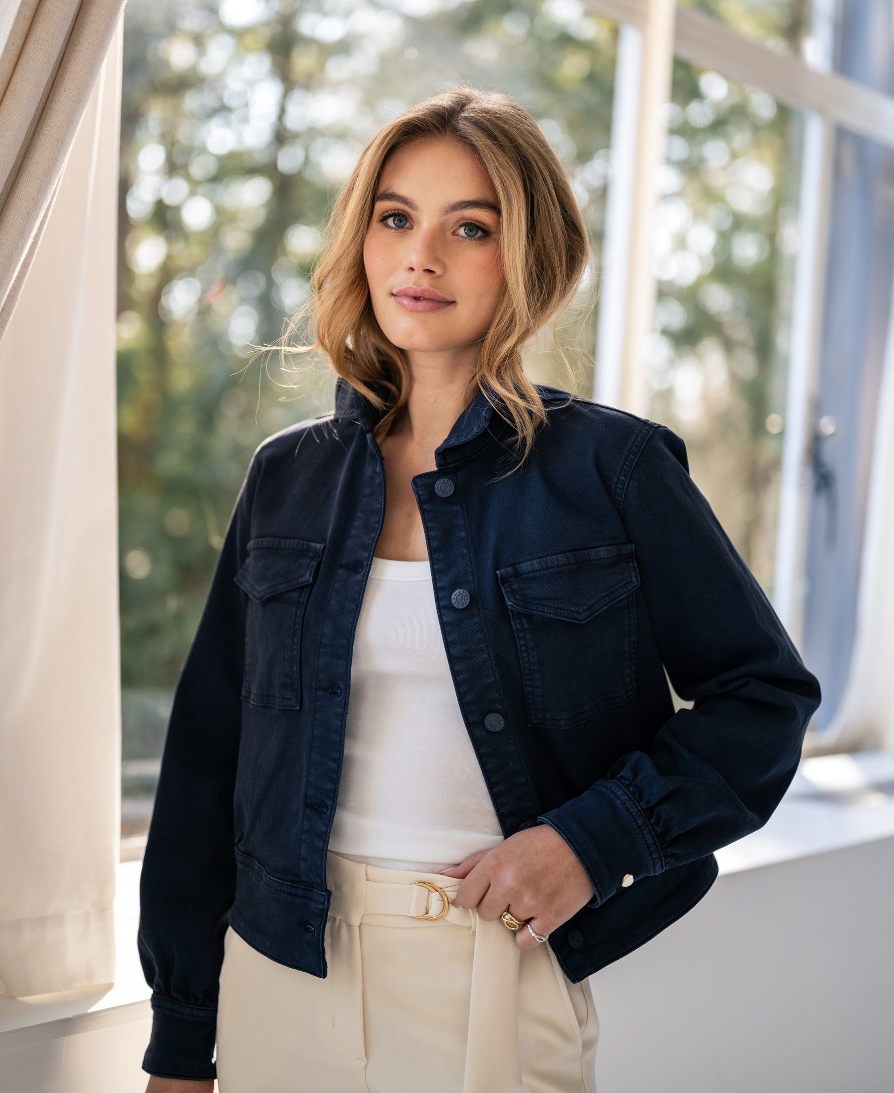 Woman wears dark blue PLEIN PUBLIQUE stretch denim jacket LA CALLAS, standing by a window.