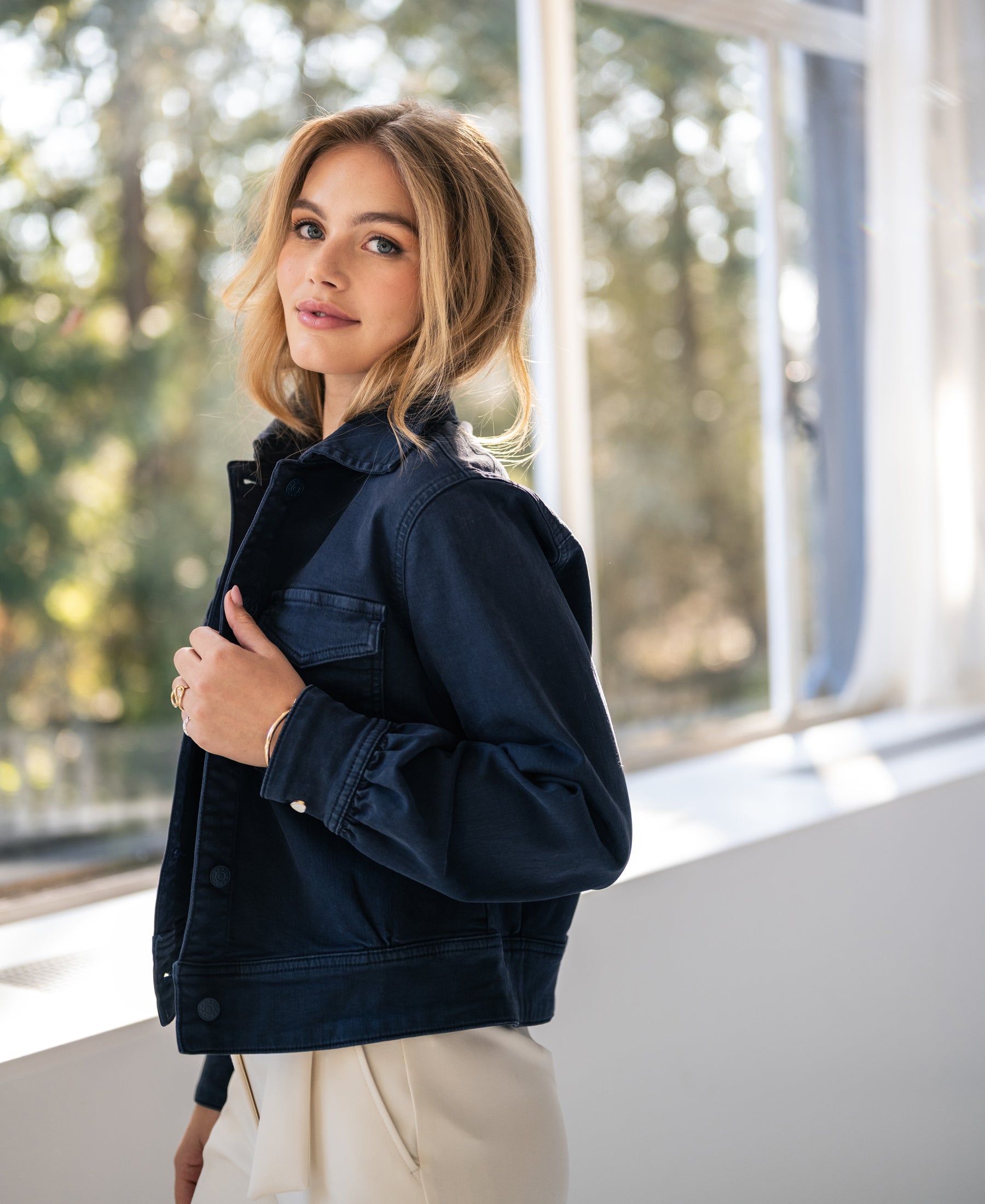 Woman wearing a PLEIN PUBLIQUE dark blue stretch denim jacket stands by a sunlit window.