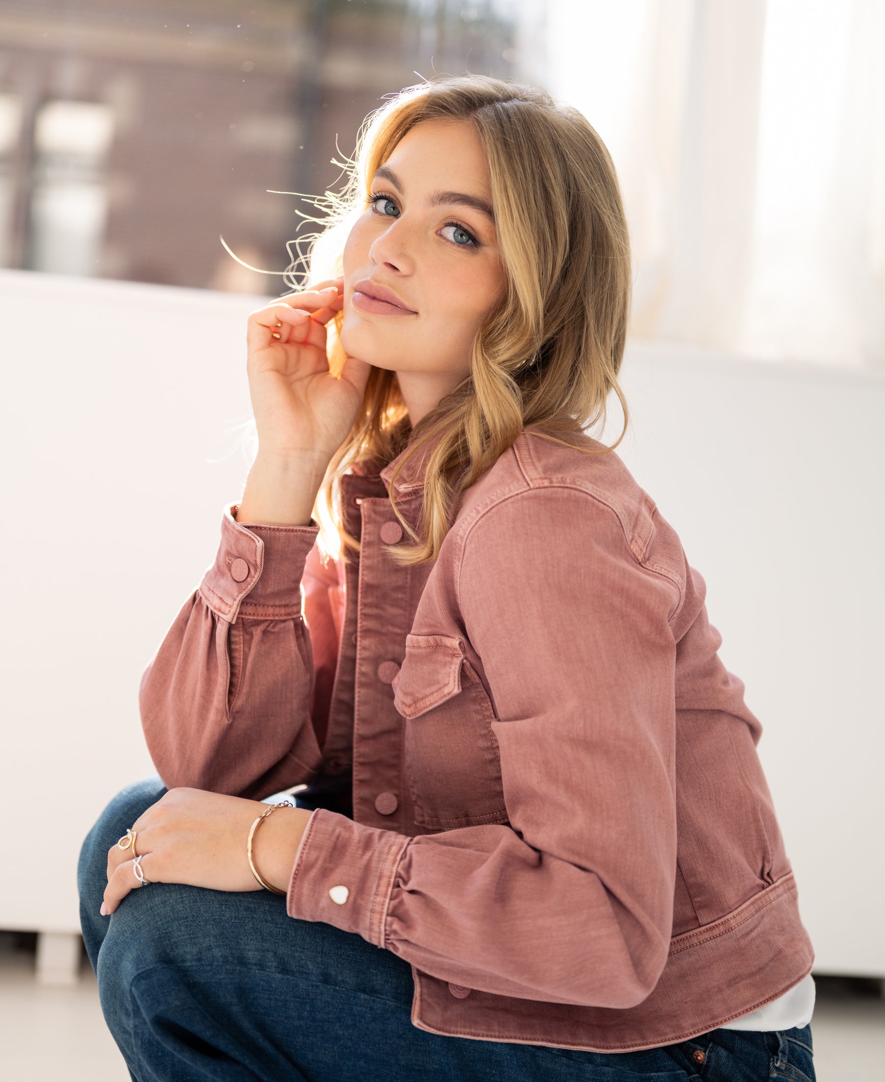 A woman with wavy blonde hair wears a stretch denim jacket in dusty pink, smiling indoors.