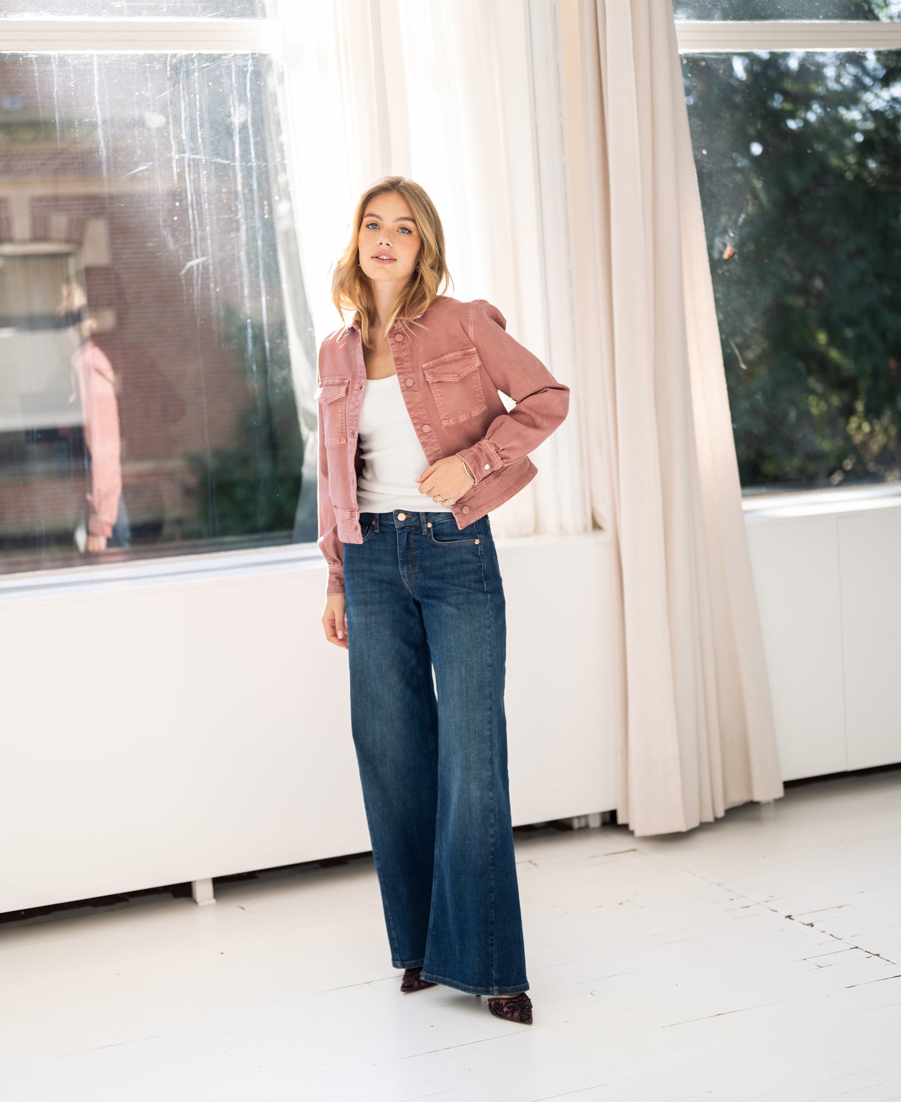 Woman stands indoors by large windows wearing a dusty pink stretch denim jacket.