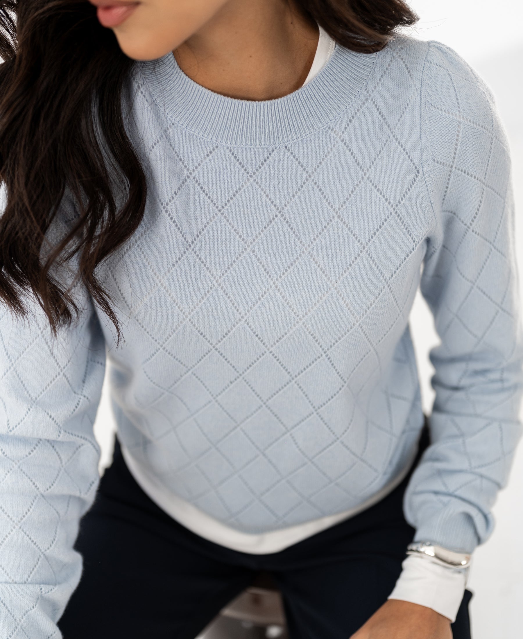 Woman wearing the PLEIN PUBLIQUE Argyle merino sweater LE CAMBRIDGE in light blue, seated sideways.