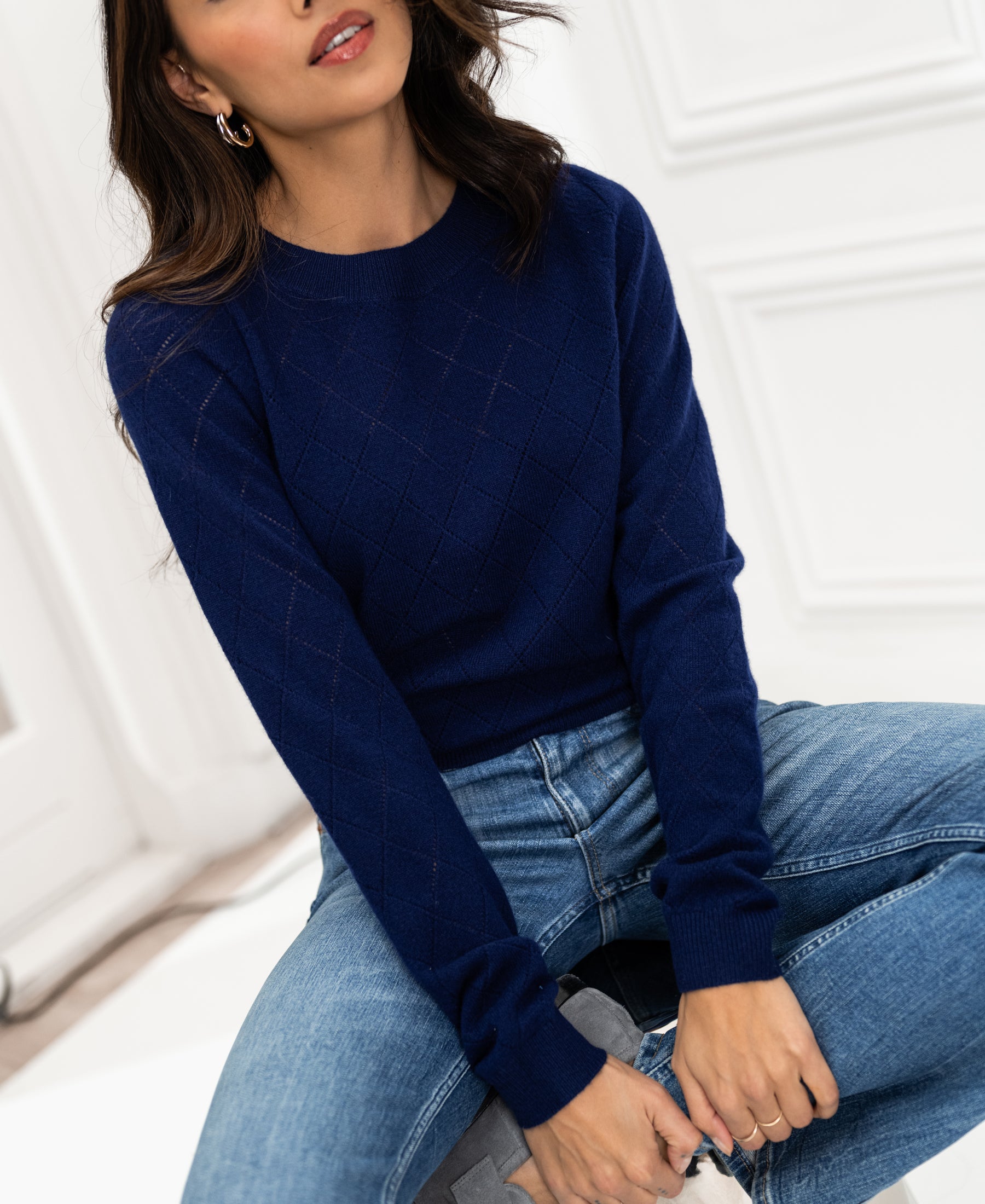 Woman wearing the PLEIN PUBLIQUE navy argyle merino wool sweater, sitting indoors with hands on her knees.