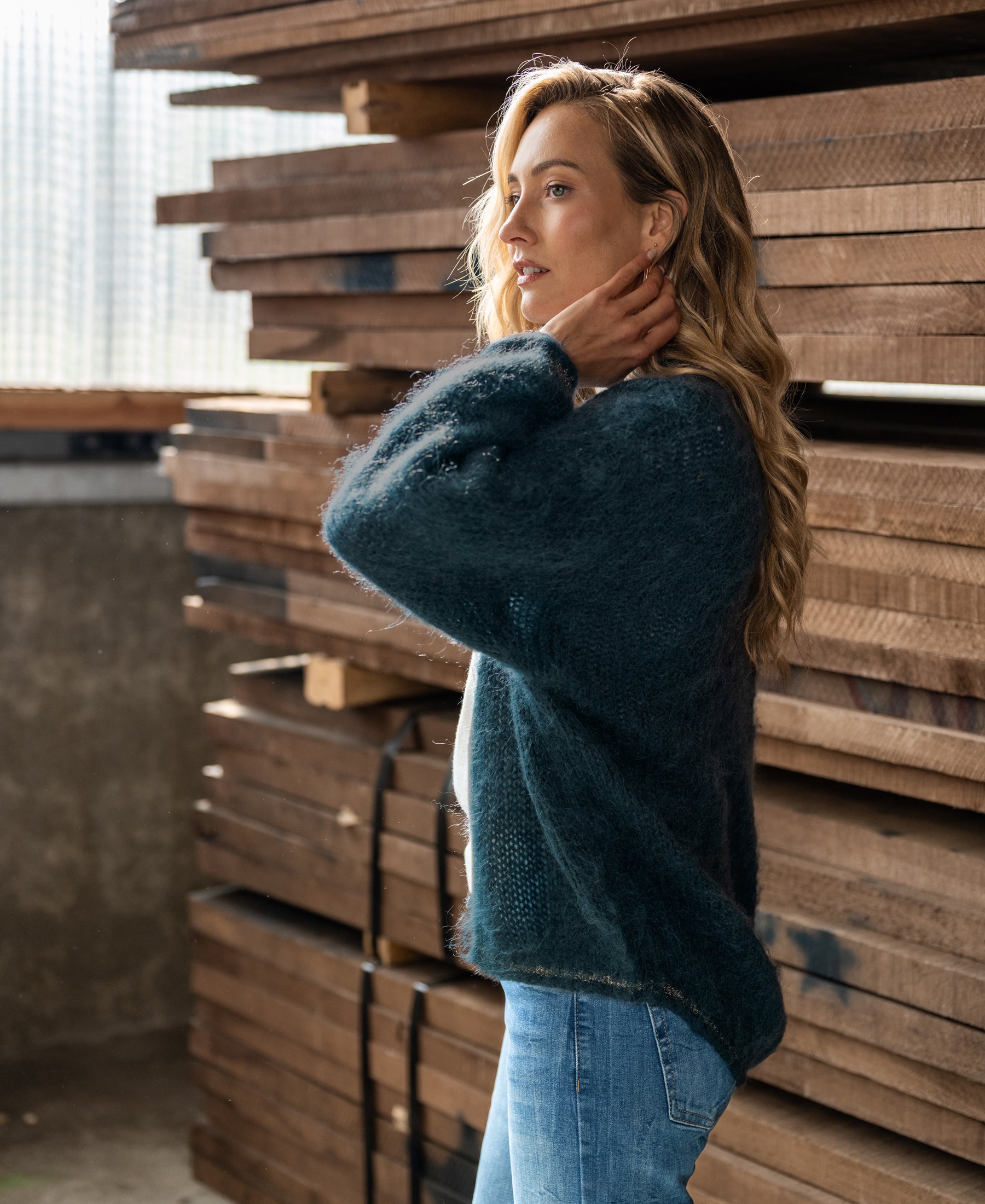 Woman wearing a fluffy dark misty green vest stands by stacked wooden planks.