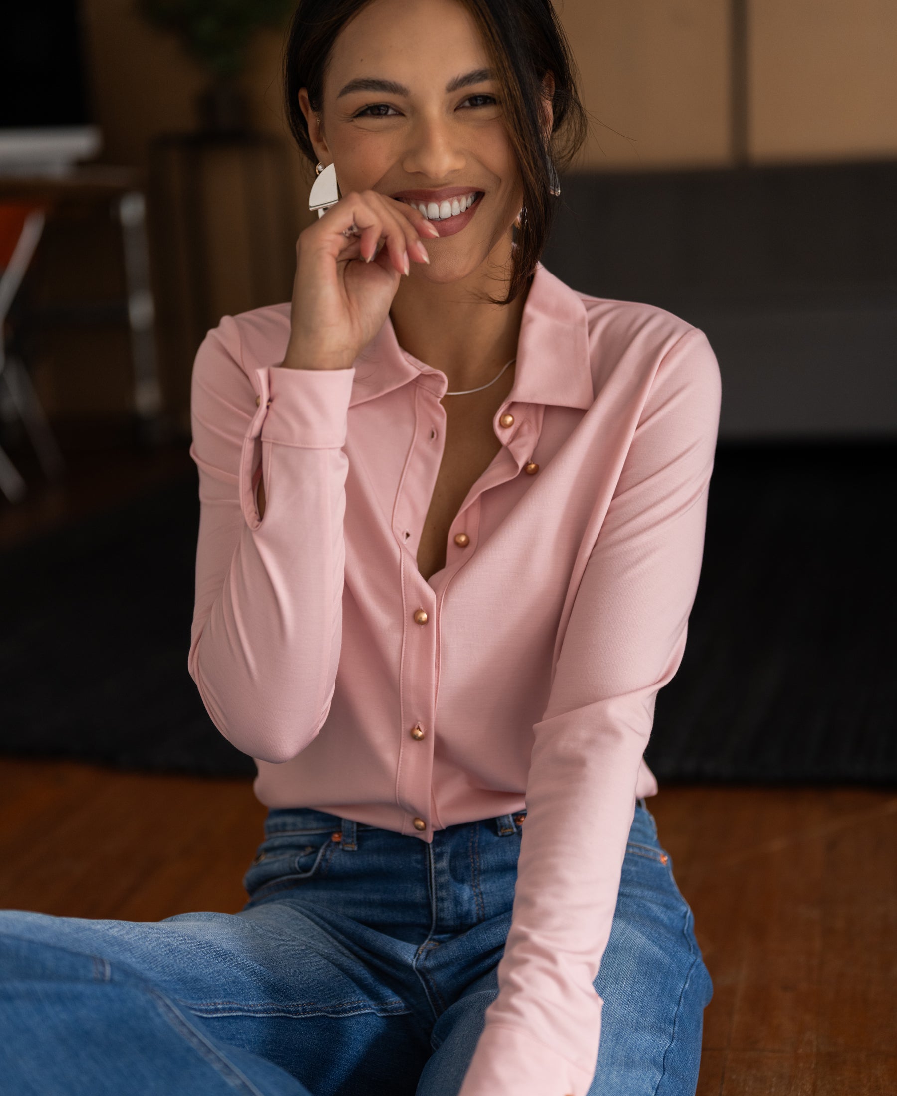 Woman indoors, smiling in PLEIN PUBLIQUE's pink Travel blouse LA CHERISE, hand on her face.