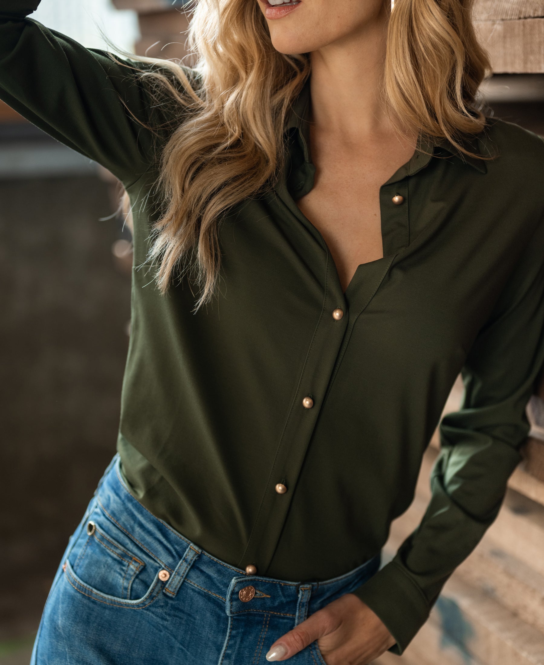 Woman wearing the PLEIN PUBLIQUE Travel blouse in army green, shown near a wooden wall.