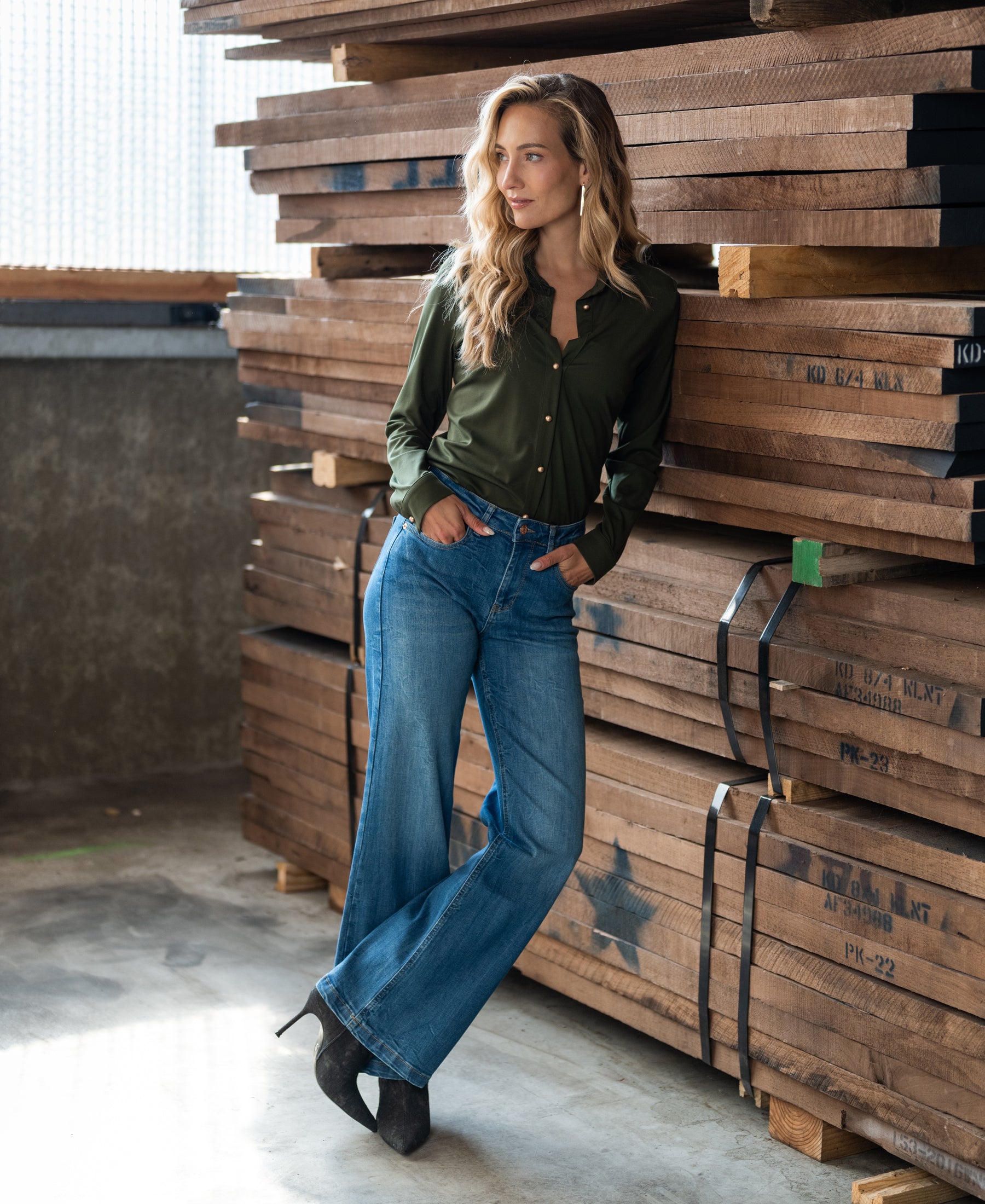 Woman wearing an army green travel blouse with gold buttons leans on stacked planks.