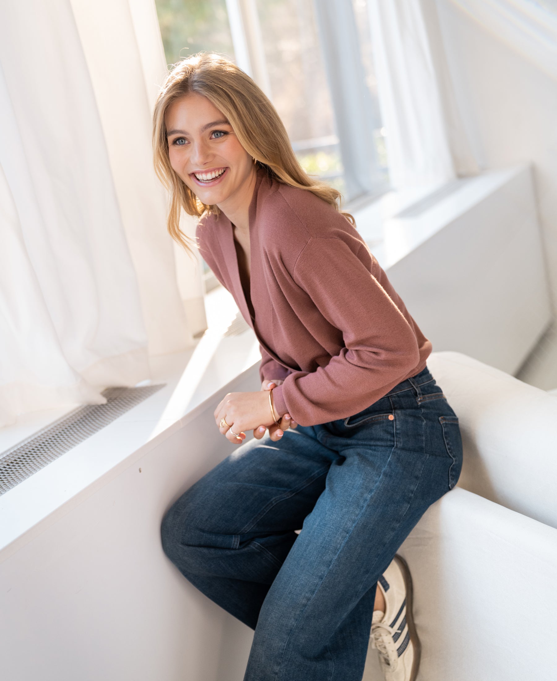Woman wearing a PLEIN PUBLIQUE wrap sweater in desert rose sits by a window, leaning on a white couch.