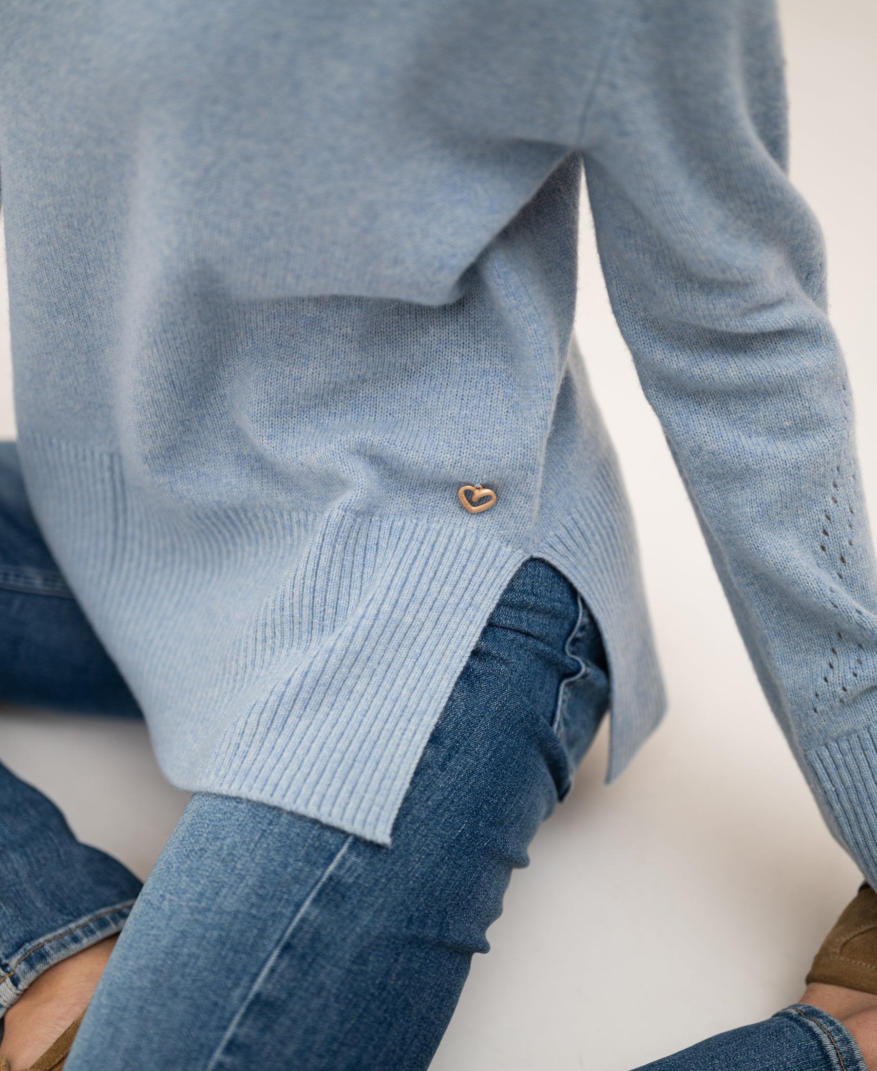 Person wears a PLEIN PUBLIQUE LA COZY light blue turtleneck sweater with a heart pin, sitting on a light surface.