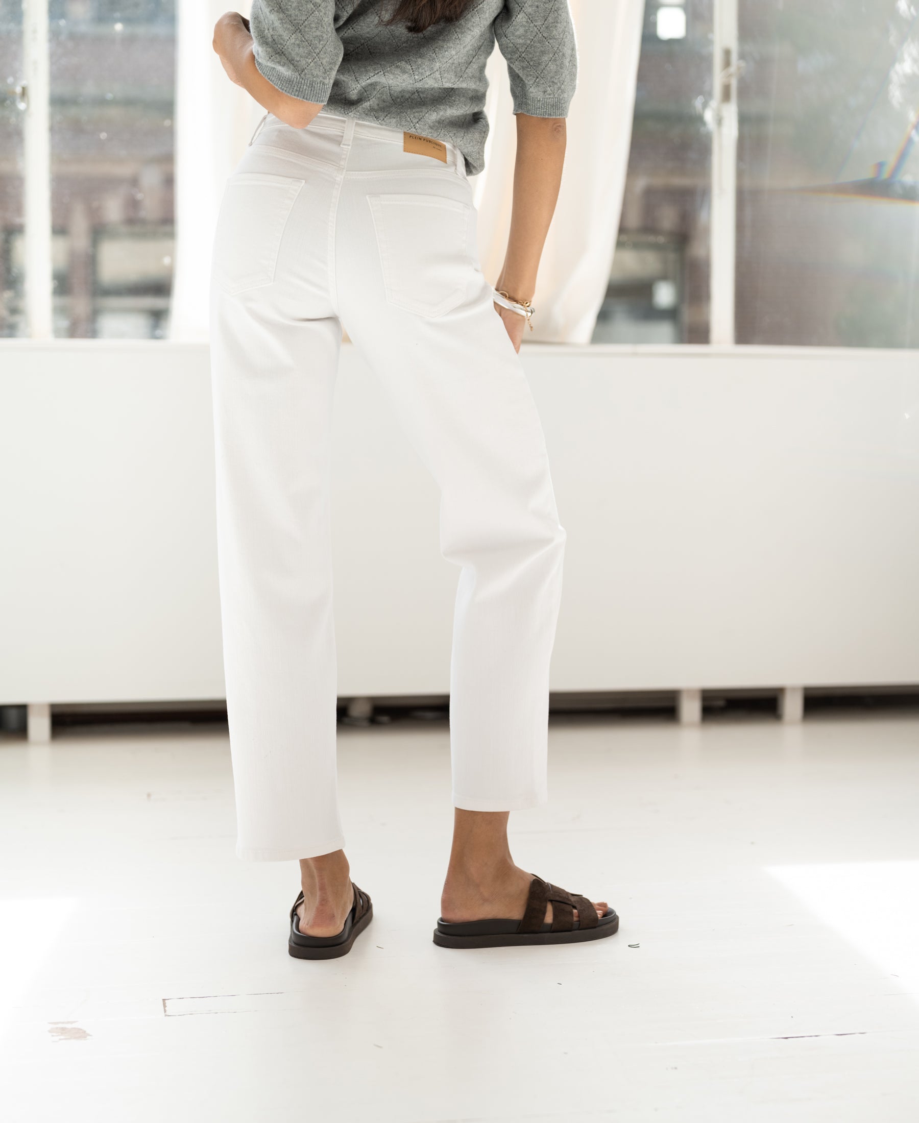Someone stands indoors facing away, wearing cropped white LE DEAN jeans.