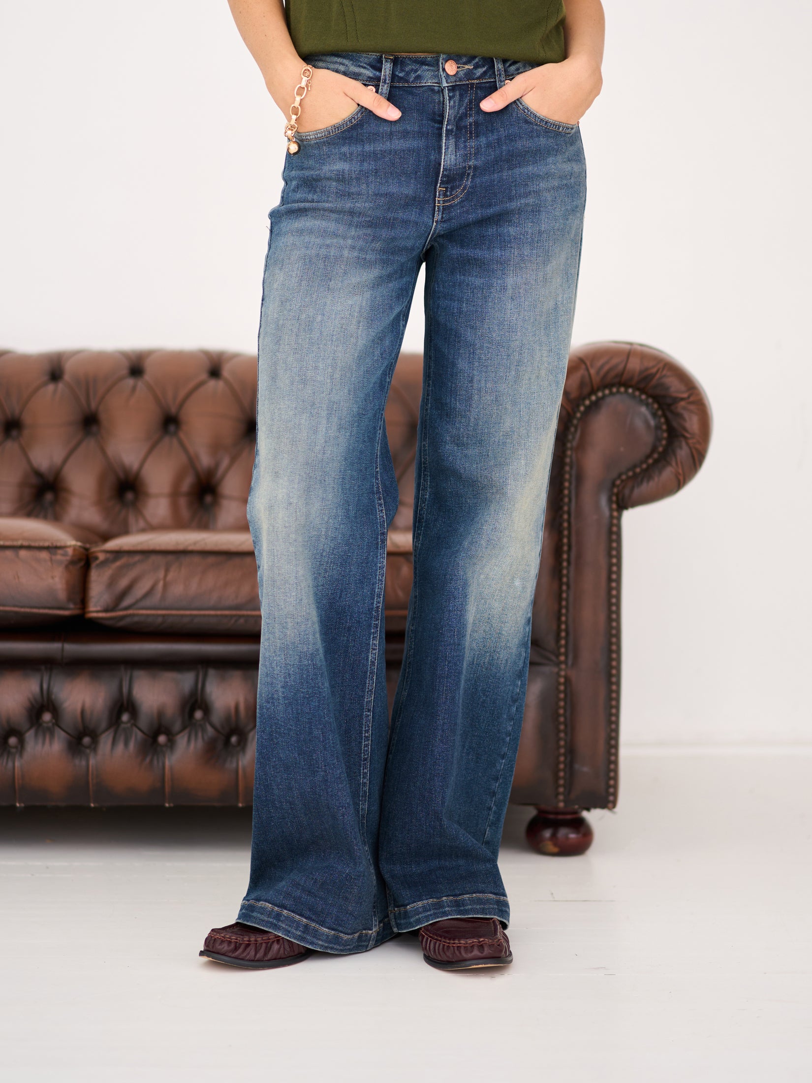 Person wearing blue vintage wide leg jeans, standing by a brown leather couch.