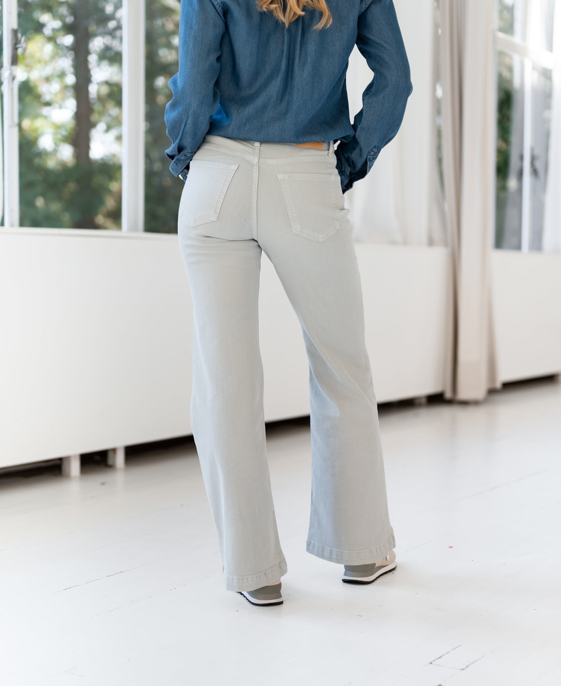 Person wearing light grey PLEIN PUBLIQUE LES DEUX jeans stands indoors, facing away by large windows.