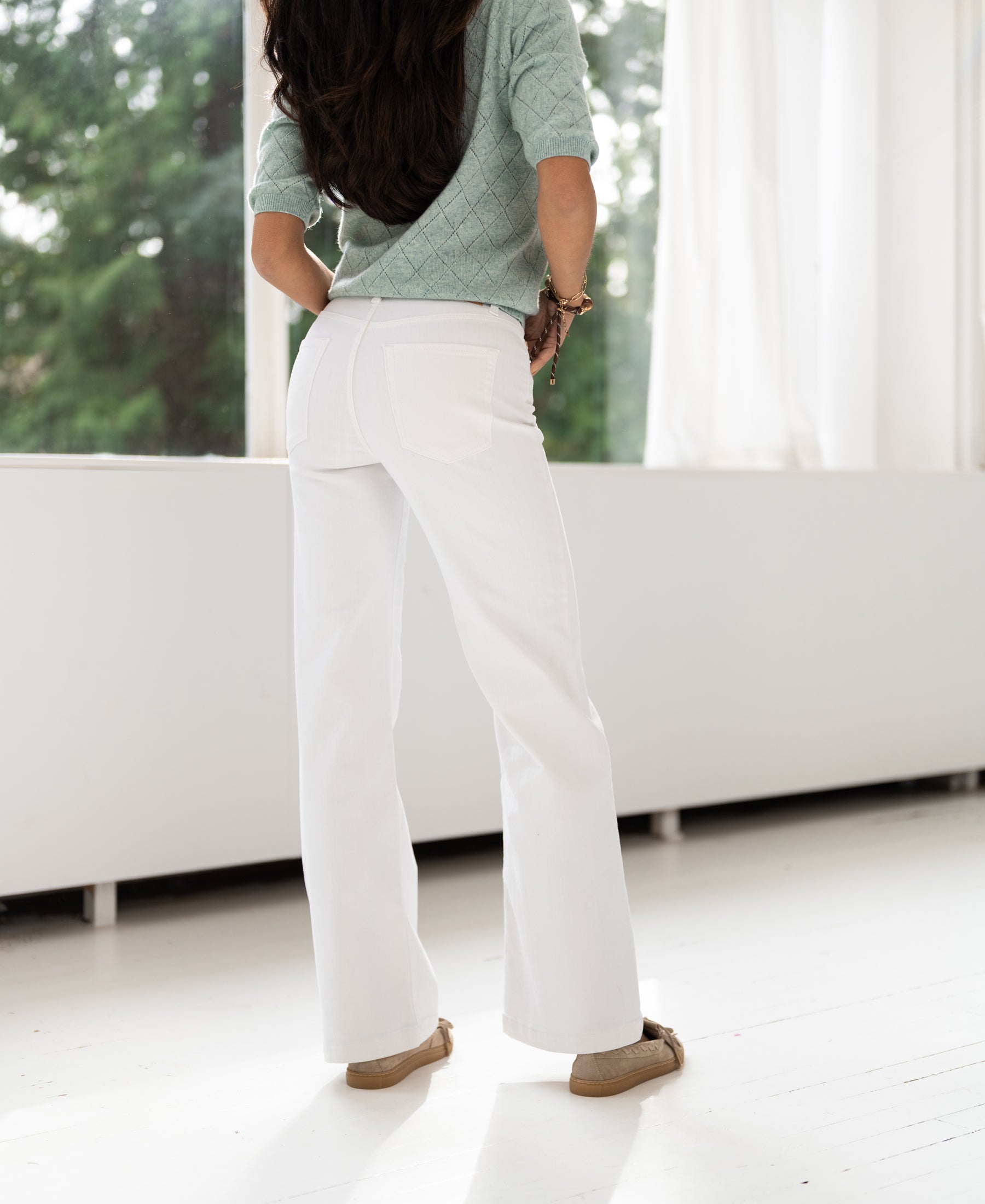 Woman standing indoors facing window, wearing wide leg jeans in white.