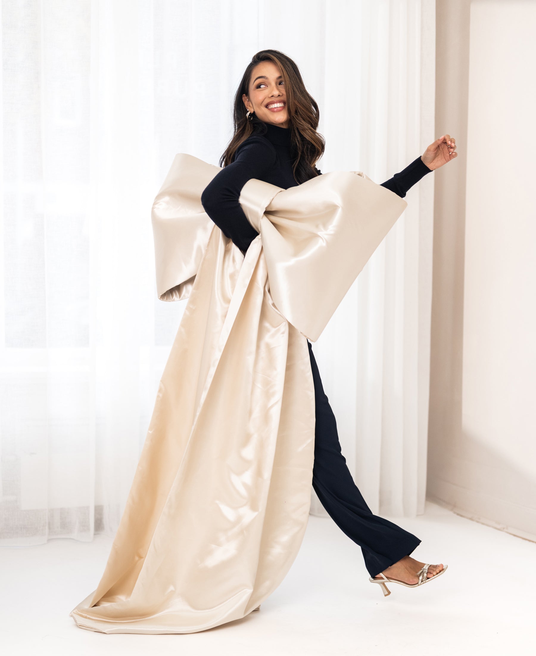 Woman wearing black PLEIN PUBLIQUE Merino wool turtleneck sweater, standing by white curtains.