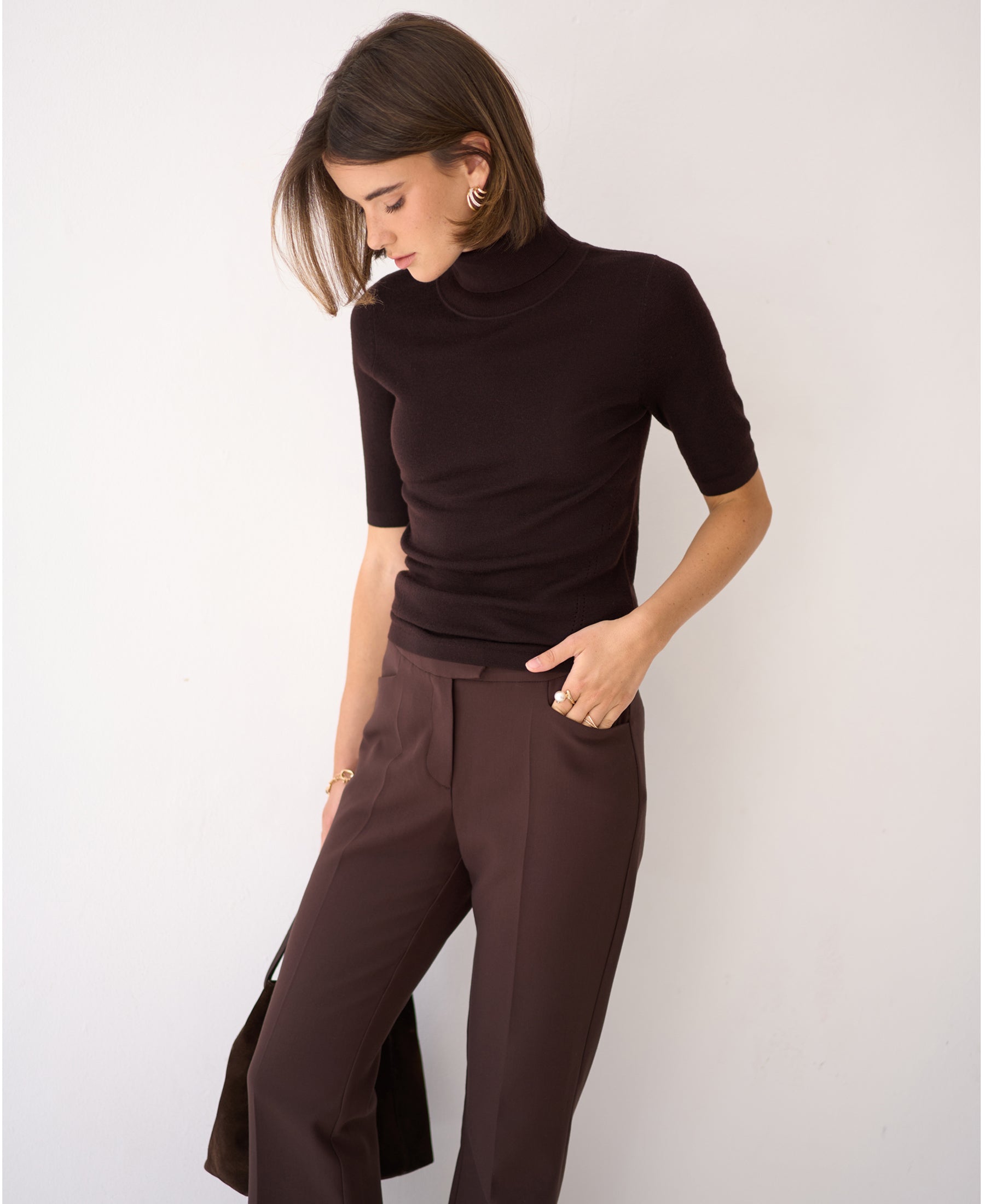 Woman in a short sleeve turtleneck sweater LE FRANCOIS Pure Choco stands by a white wall, looking down.