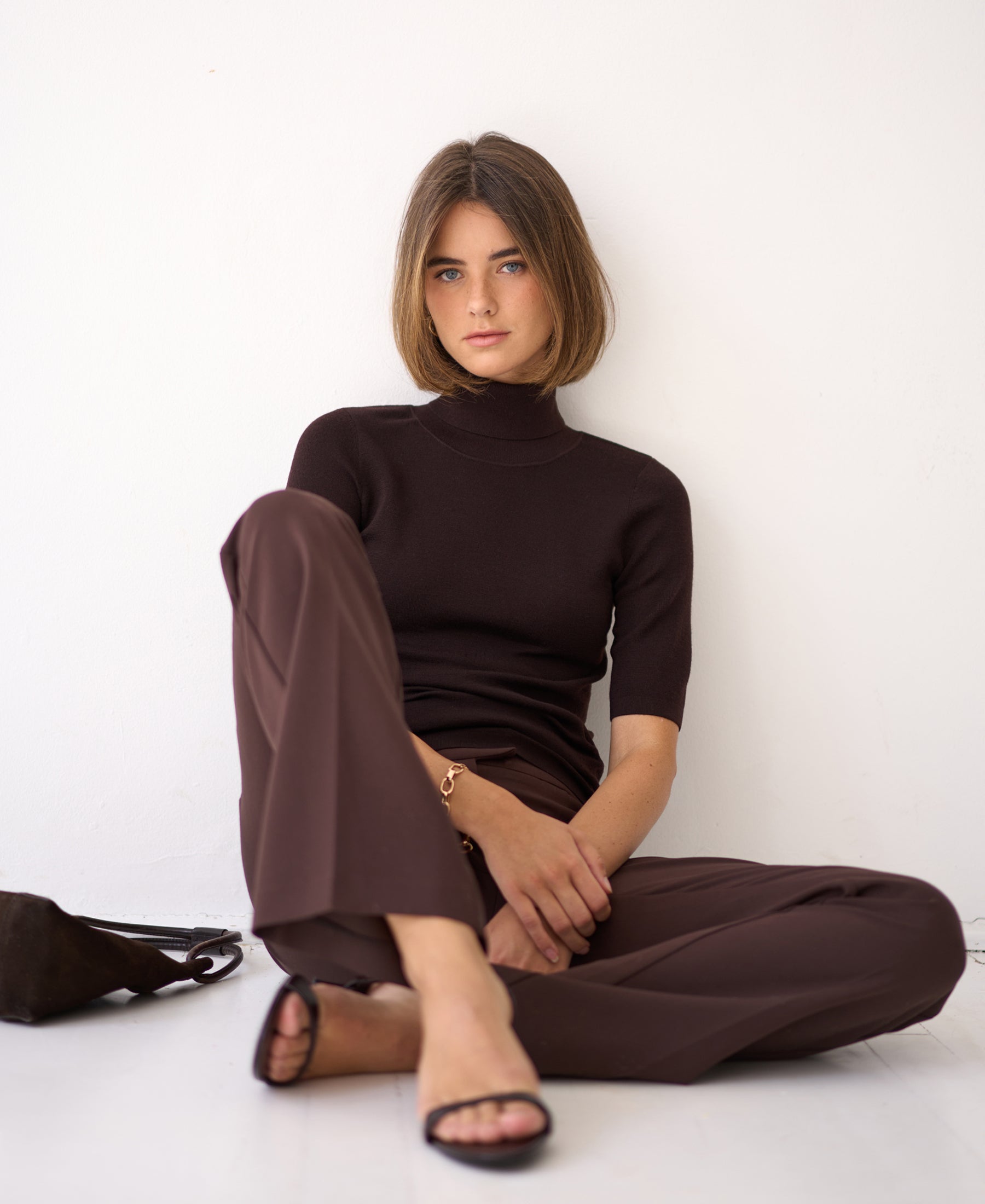 Brown short-sleeved turtleneck sweater, Pure Choco color, shown on a woman seated indoors against a white wall.