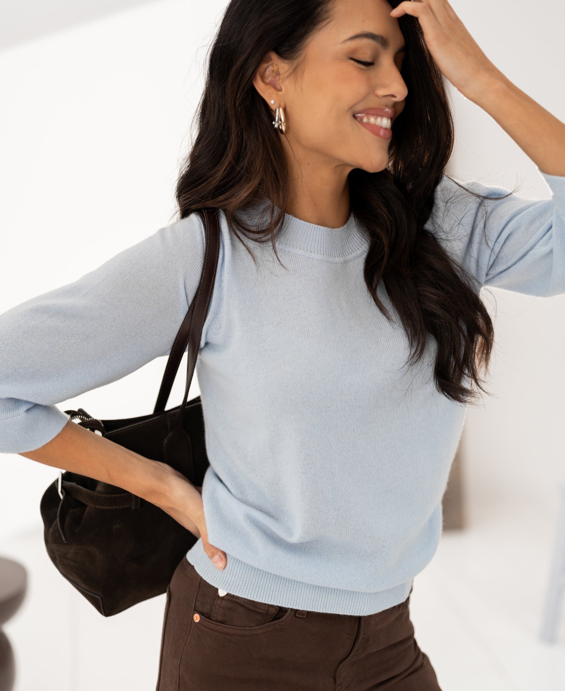 Woman smiles with closed eyes wearing the LE FREJUS Light Blue Merino Sweater by PLEIN PUBLIQUE.