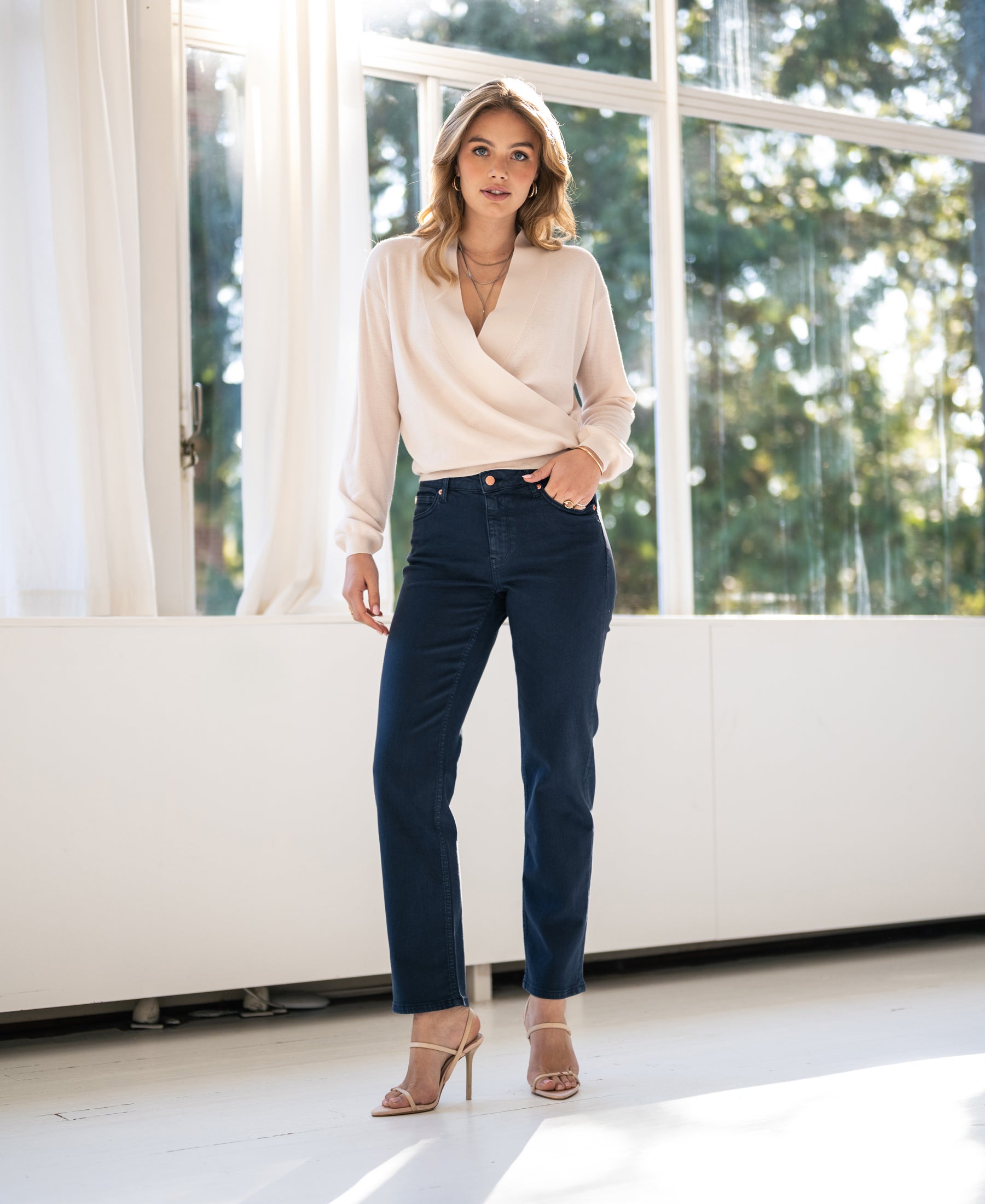 Woman indoors by large windows, wearing dark blue PLEIN PUBLIQUE cropped jeans.