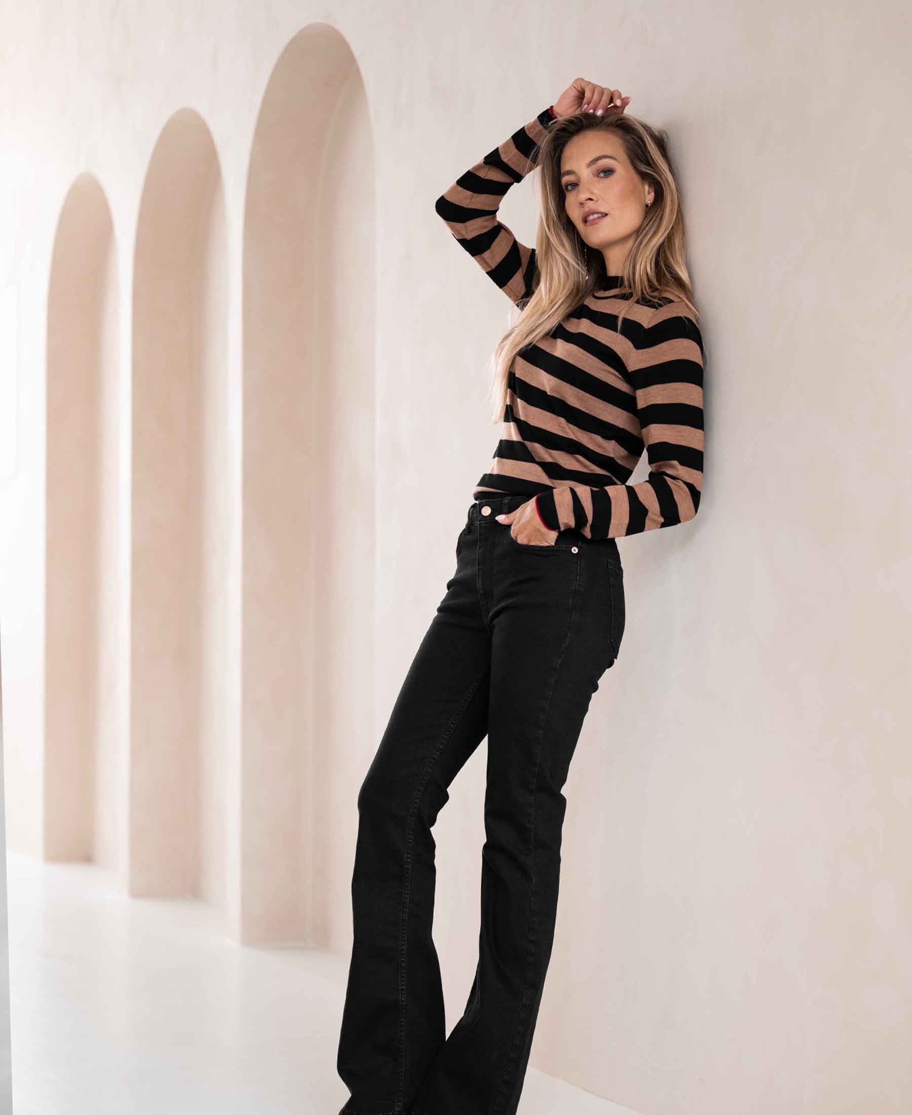 Woman wearing the LE JEUDI Black-Chai Tea Striped Turtleneck sweater, leaning against a wall with arched alcoves.
