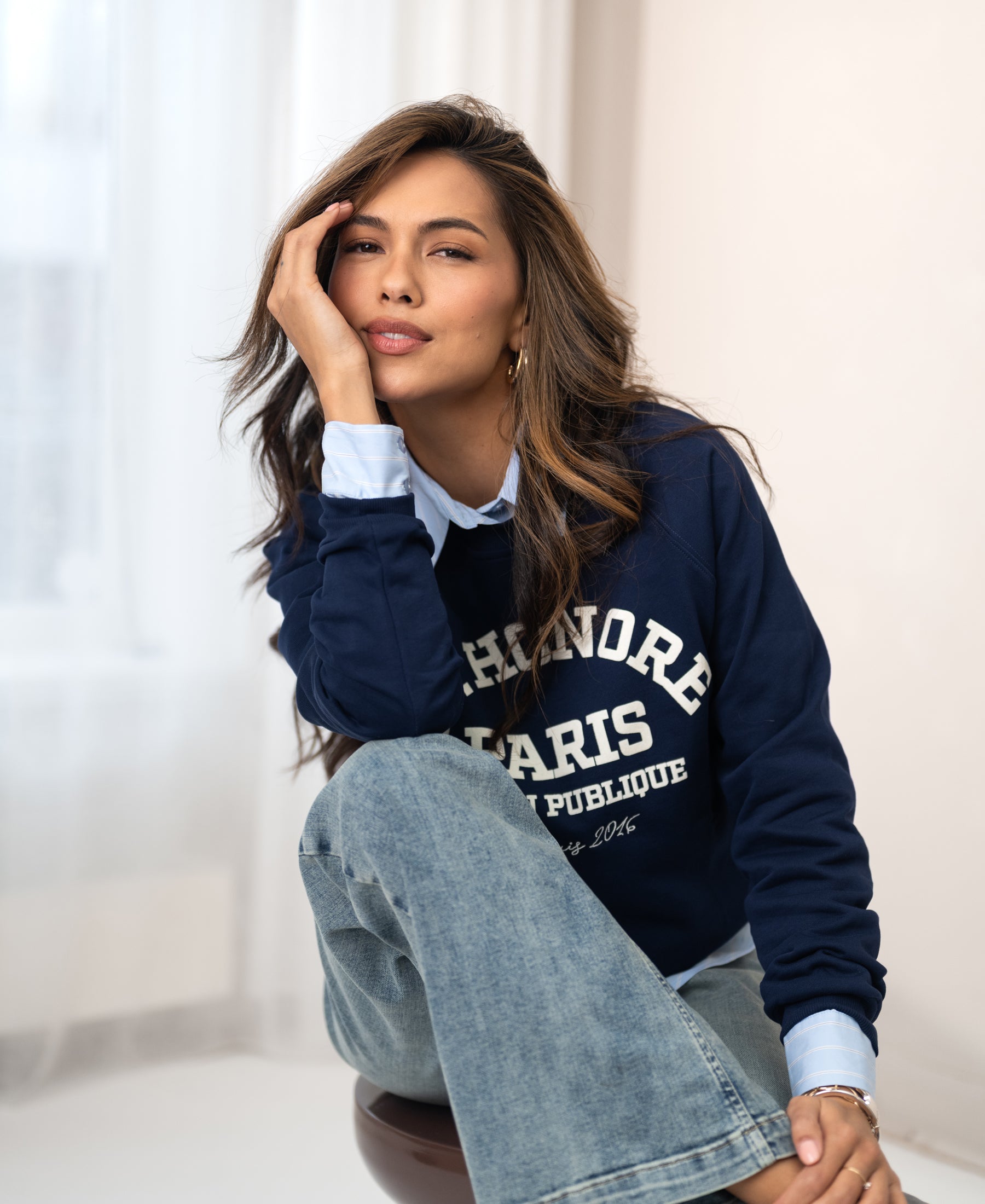 Woman indoors in the LA HARPER dark blue sweater, smiling softly with her head resting on her hand.
