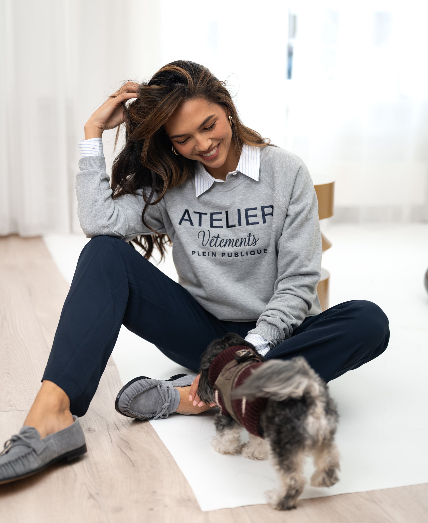 Woman wearing a light grey PLEIN PUBLIQUE sweater smiles and pets her dog.