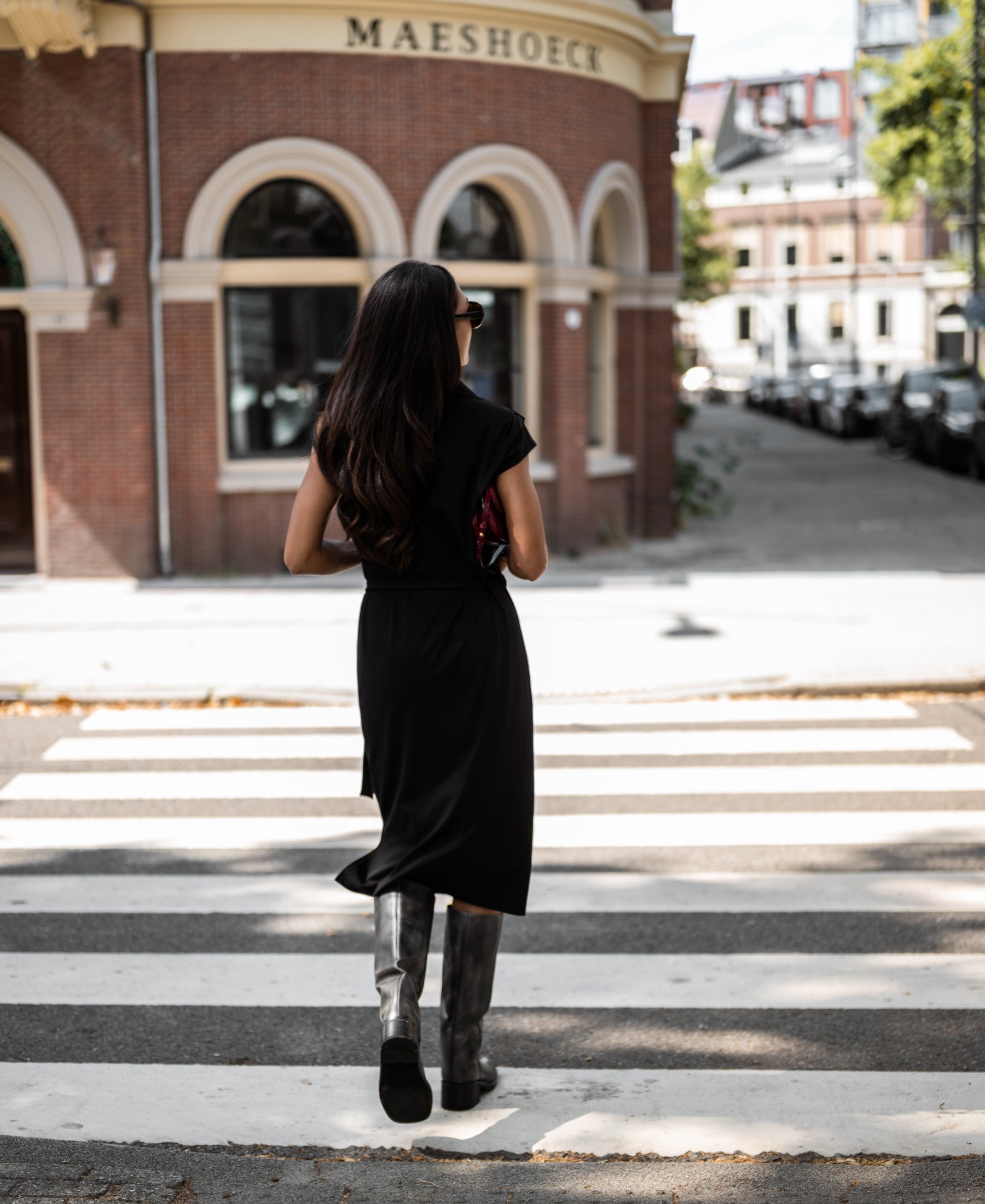 Woman wearing a black PLEIN PUBLIQUE Ines travel polo dress crossing a street with a brick building in the background.