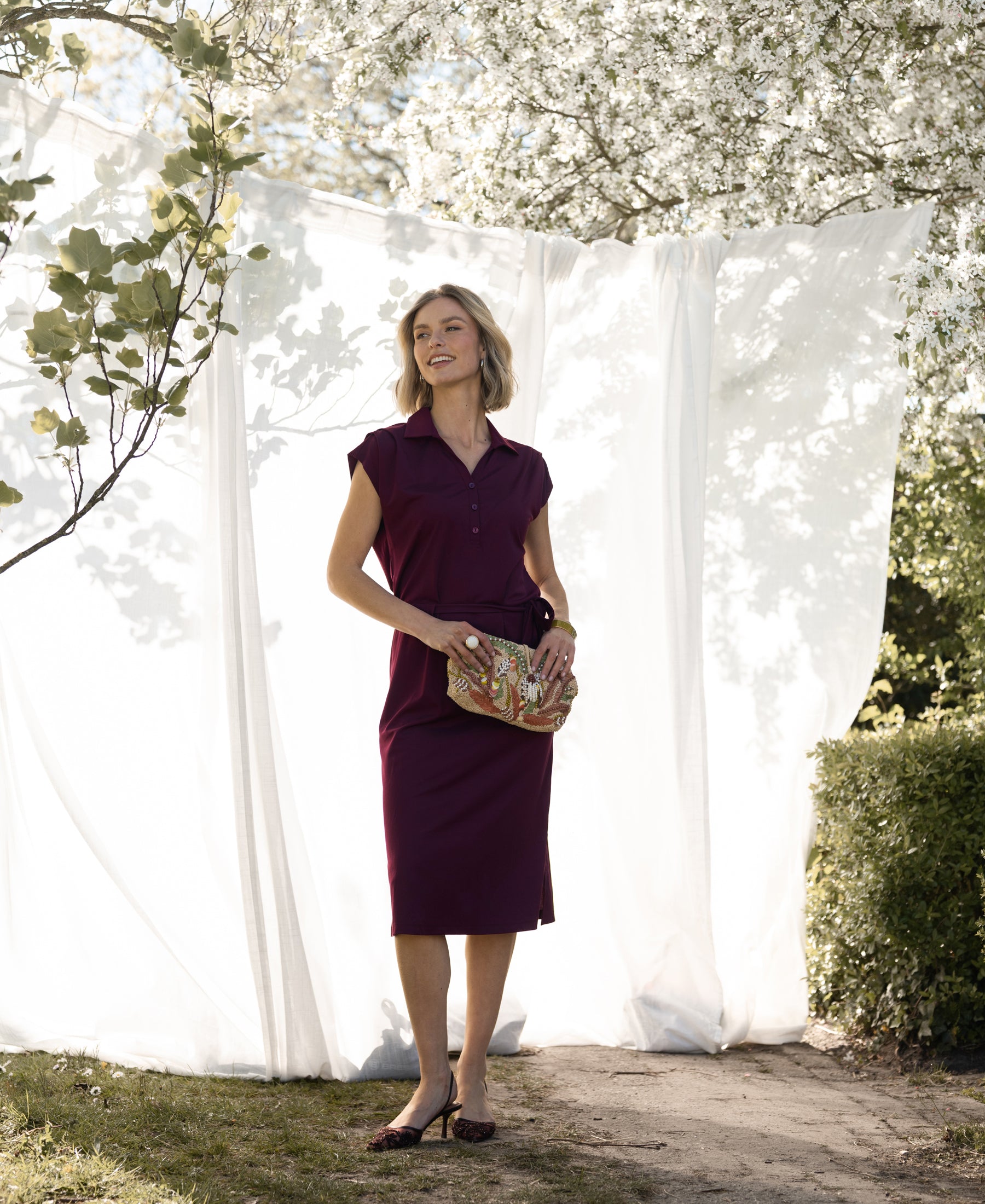 Woman in a wine-colored Travel polo dress stands outdoors with trees and white curtains in the background.