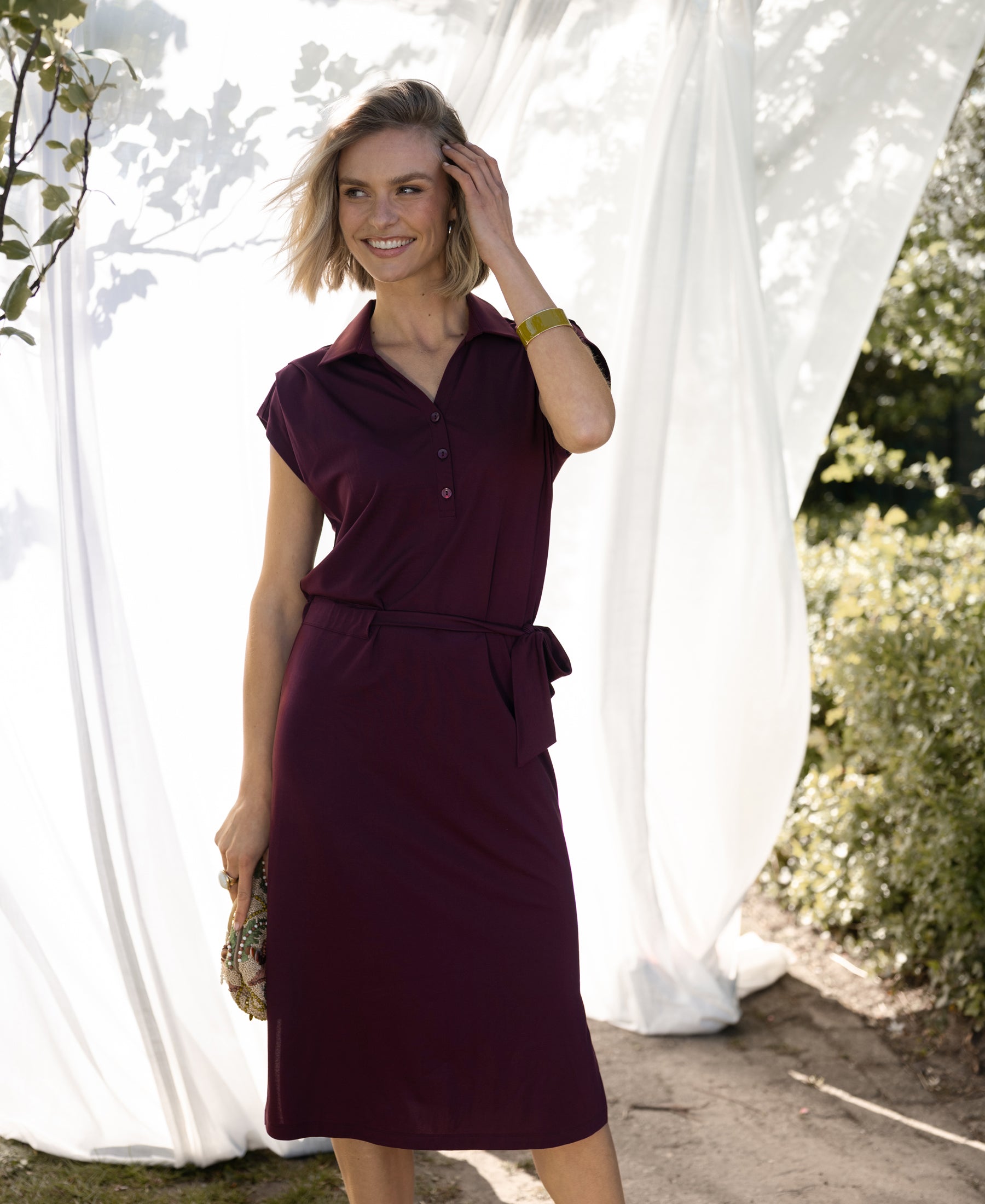 Woman wearing a travel polo dress in wine color stands outdoors by white fabric panels.