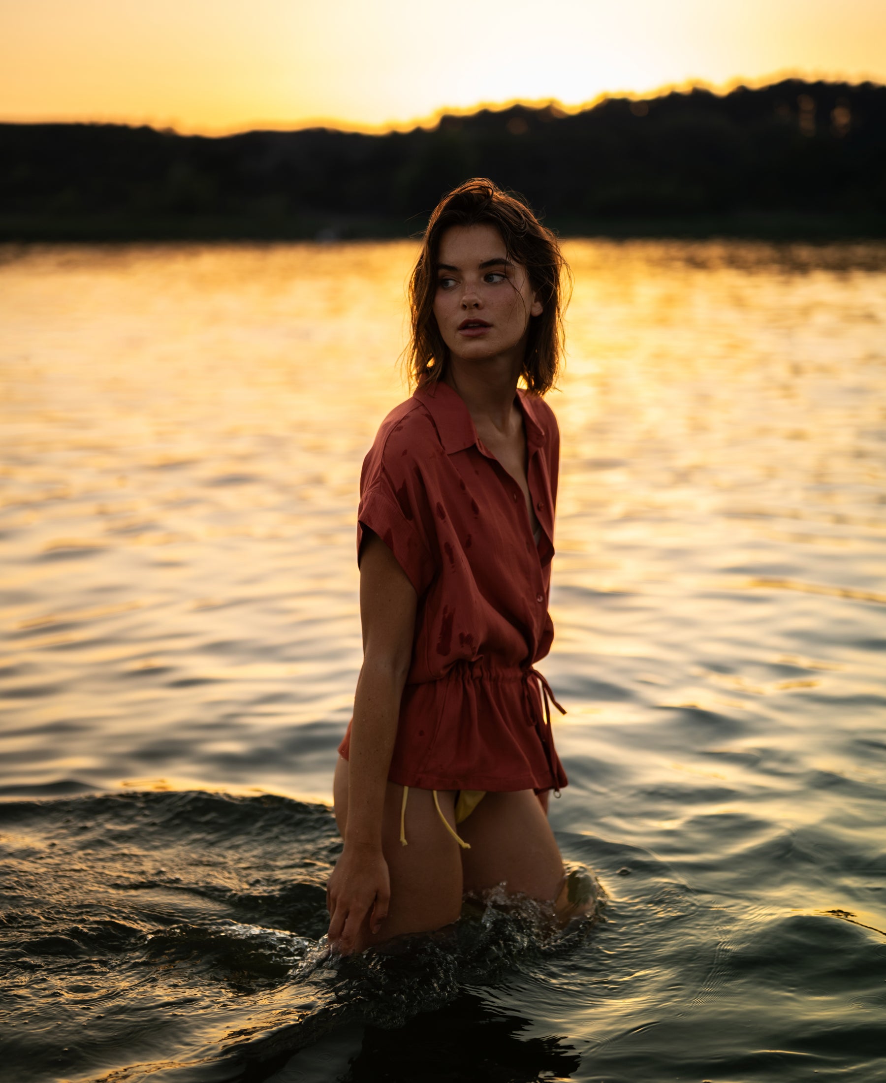 Terra-colored LA JODY blouse by PLEIN PUBLIQUE shown on model waist-deep in water at sunset, side view.