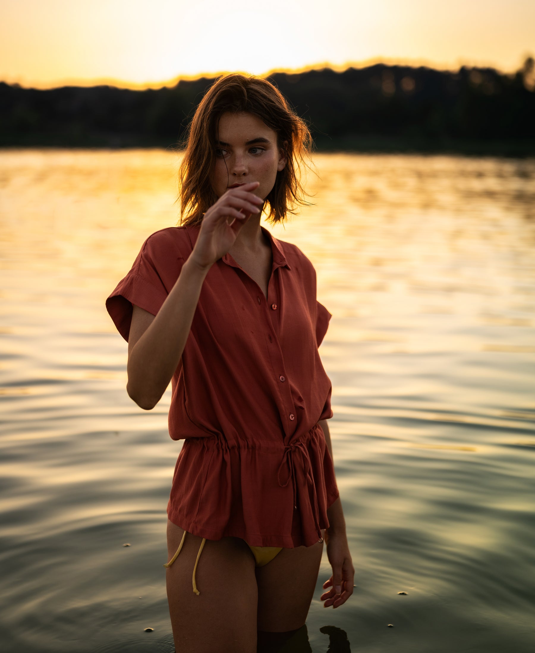 The LA JODY Terra blouse by PLEIN PUBLIQUE in terra color, shown against sunset water with a backdrop of silhouetted trees.