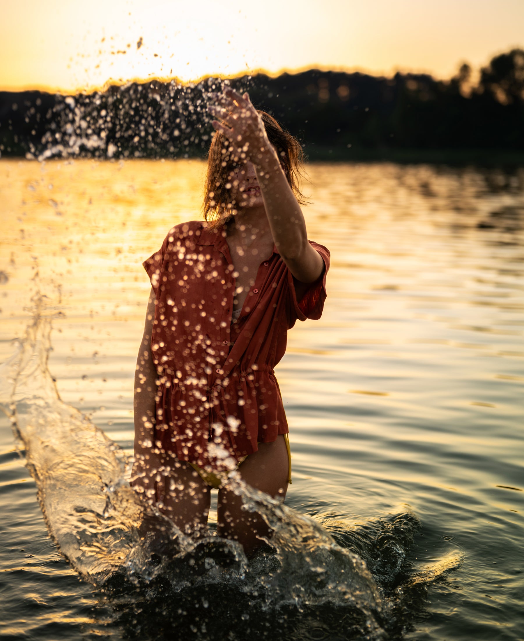 Red LA JODY Terra blouse by PLEIN PUBLIQUE shown outdoors at sunset, with water droplets in mid-air.