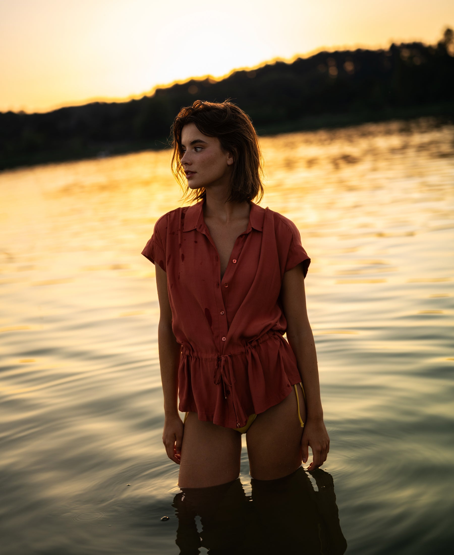 Woman wearing a Terra-colored blouse stands in water at sunset with silhouetted trees behind her.