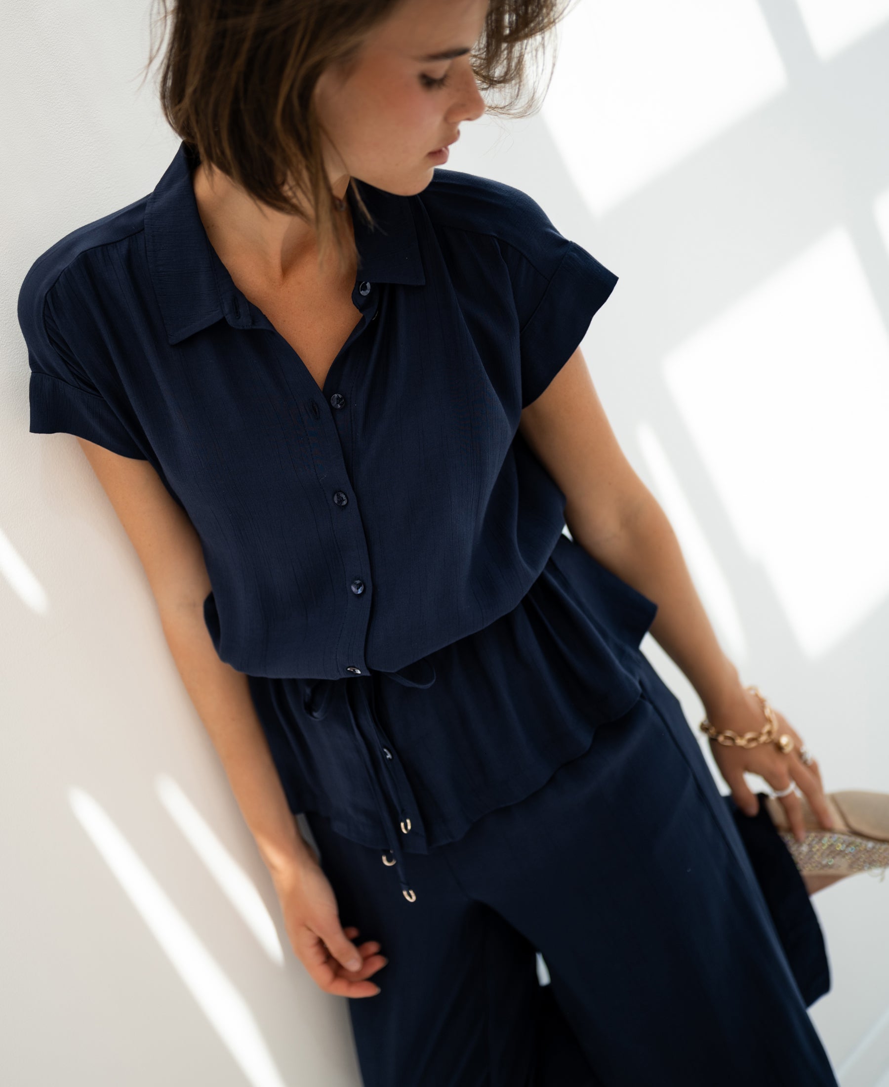 Navy blue PLEIN PUBLIQUE LA JODY blouse displayed on woman standing by a white wall, holding a glittery shoe.