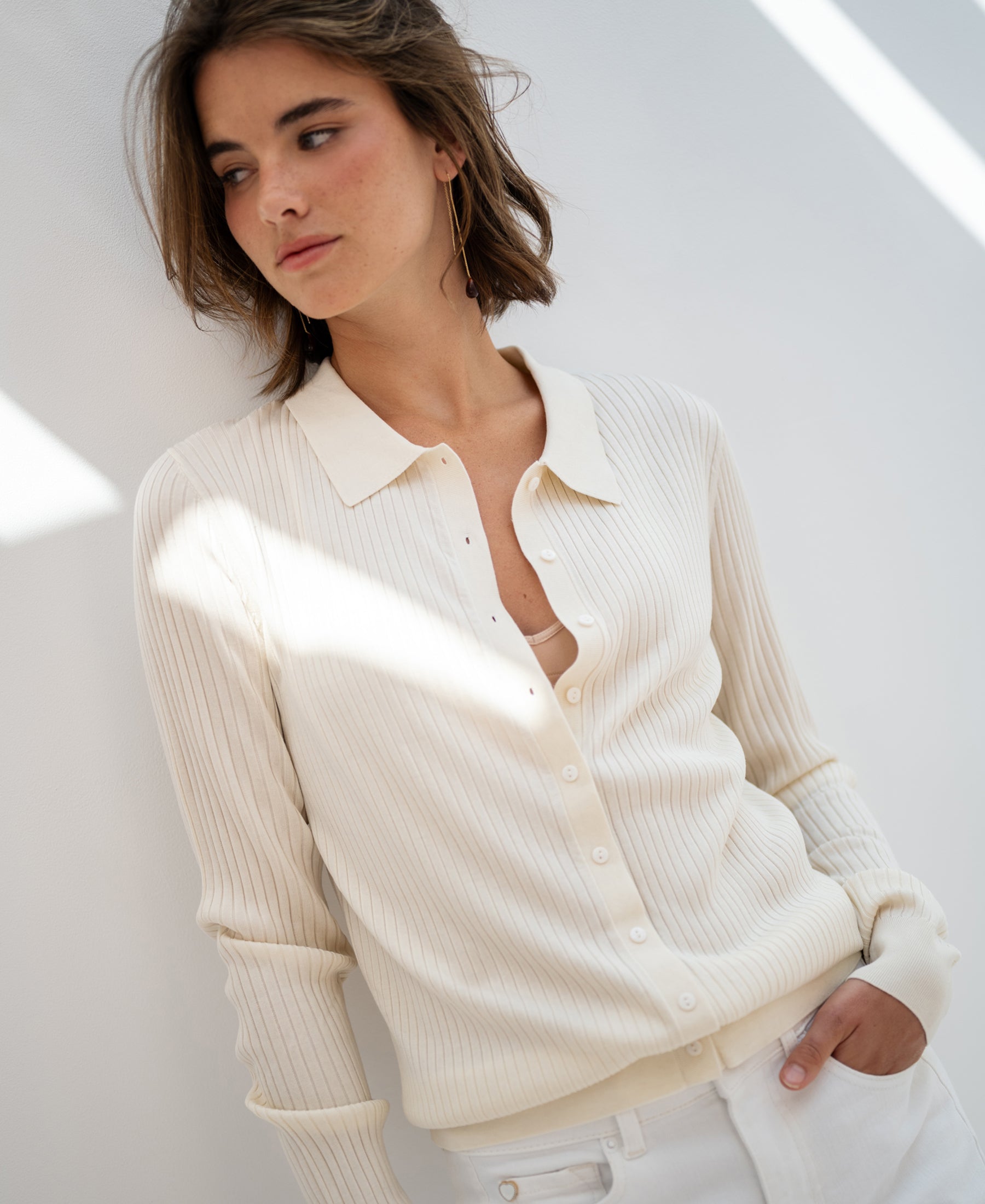 Woman wearing a cream Tencel-silk ribbed cardigan stands against a white wall, looking aside.