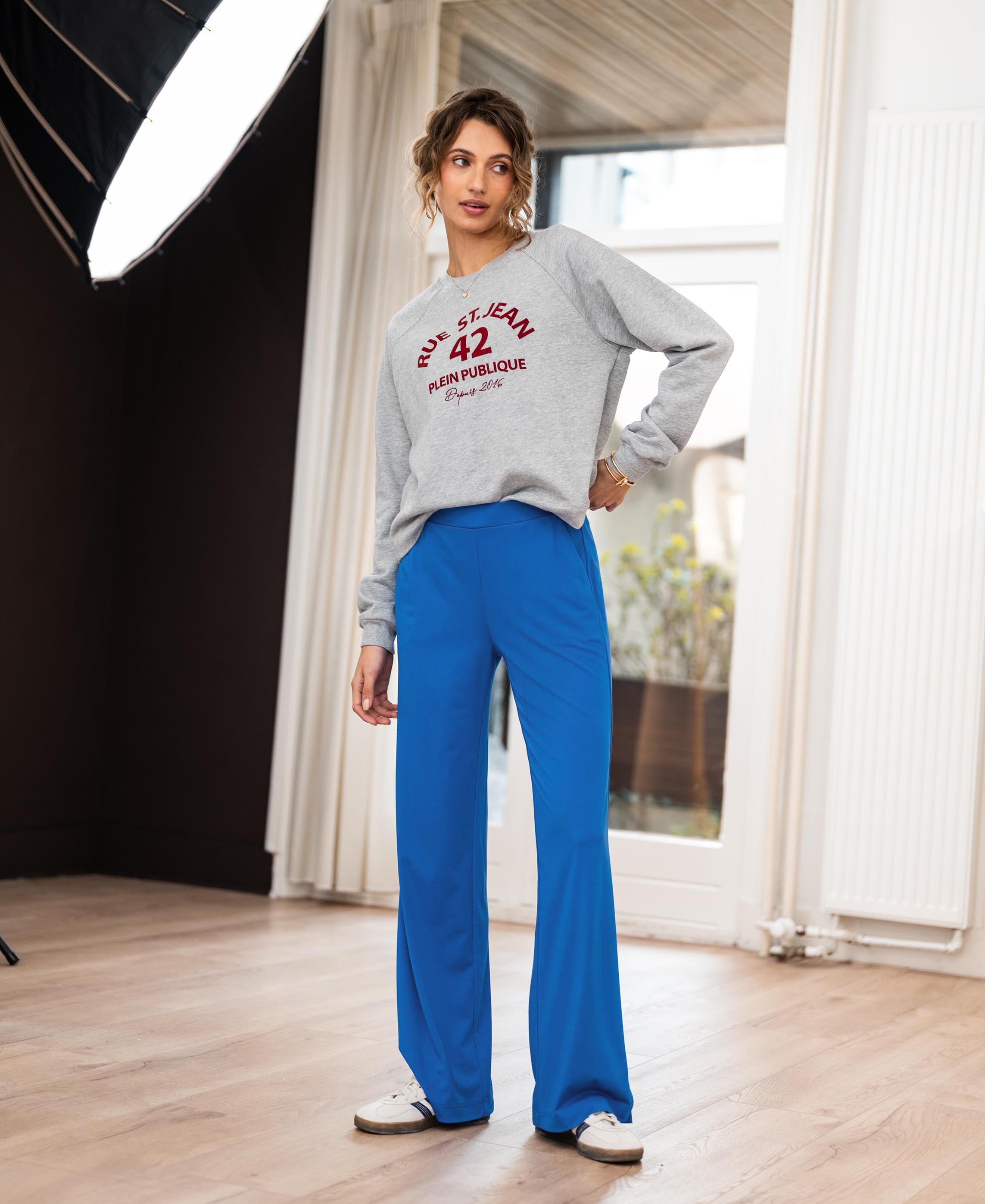 Woman indoors wearing a grey PLEIN PUBLIQUE LA SWEAT sweater on a wooden floor.