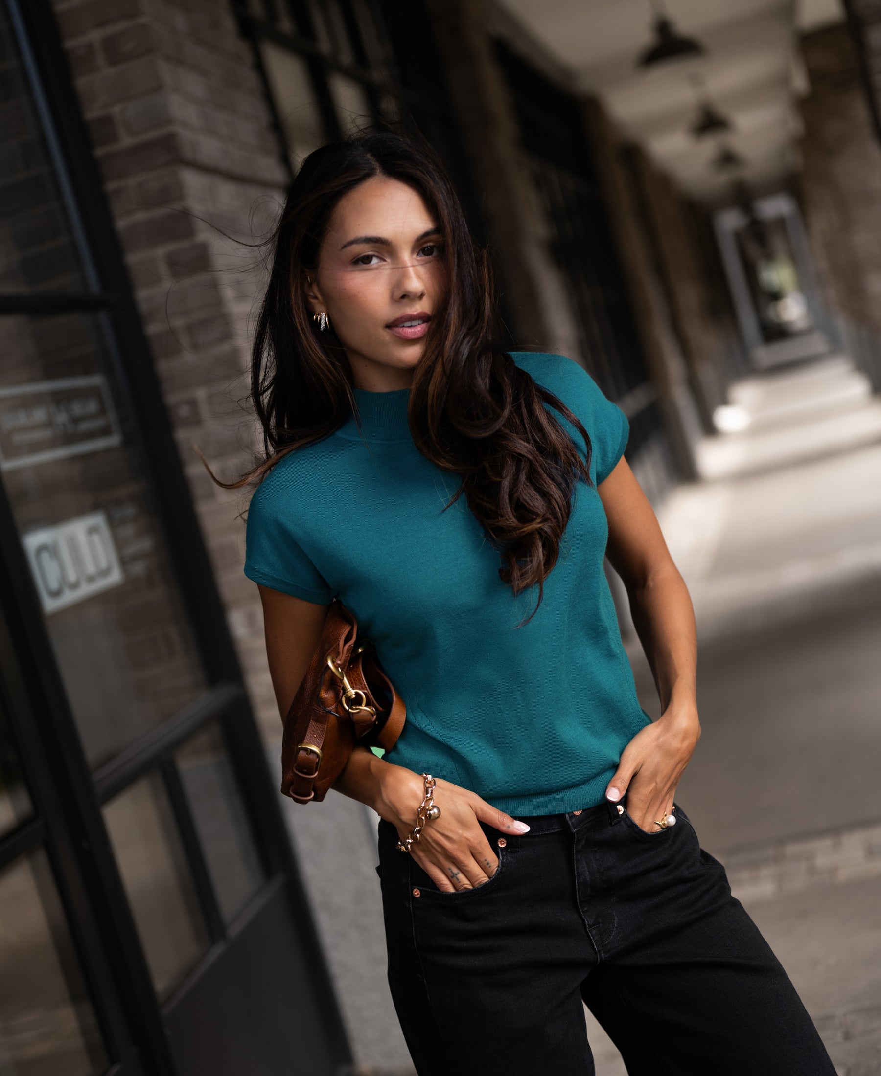 Woman wears the Merino wool LE JULIAN collared top in dark teal, standing outside a brick building.