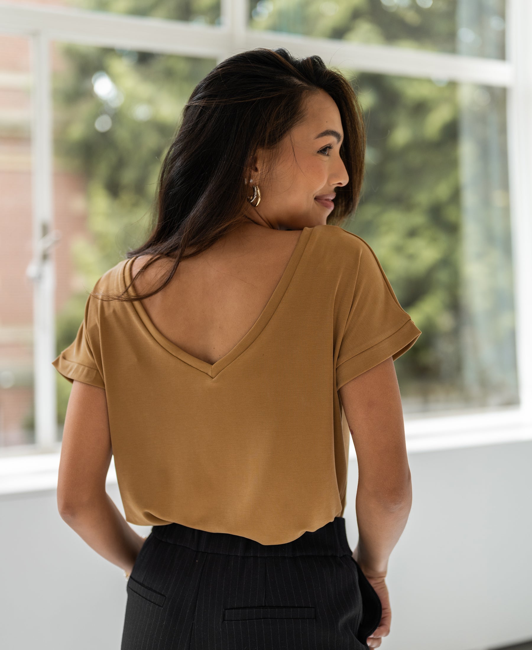 Woman with long dark hair wears light camel two-way T-shirt, shown from the back indoors.