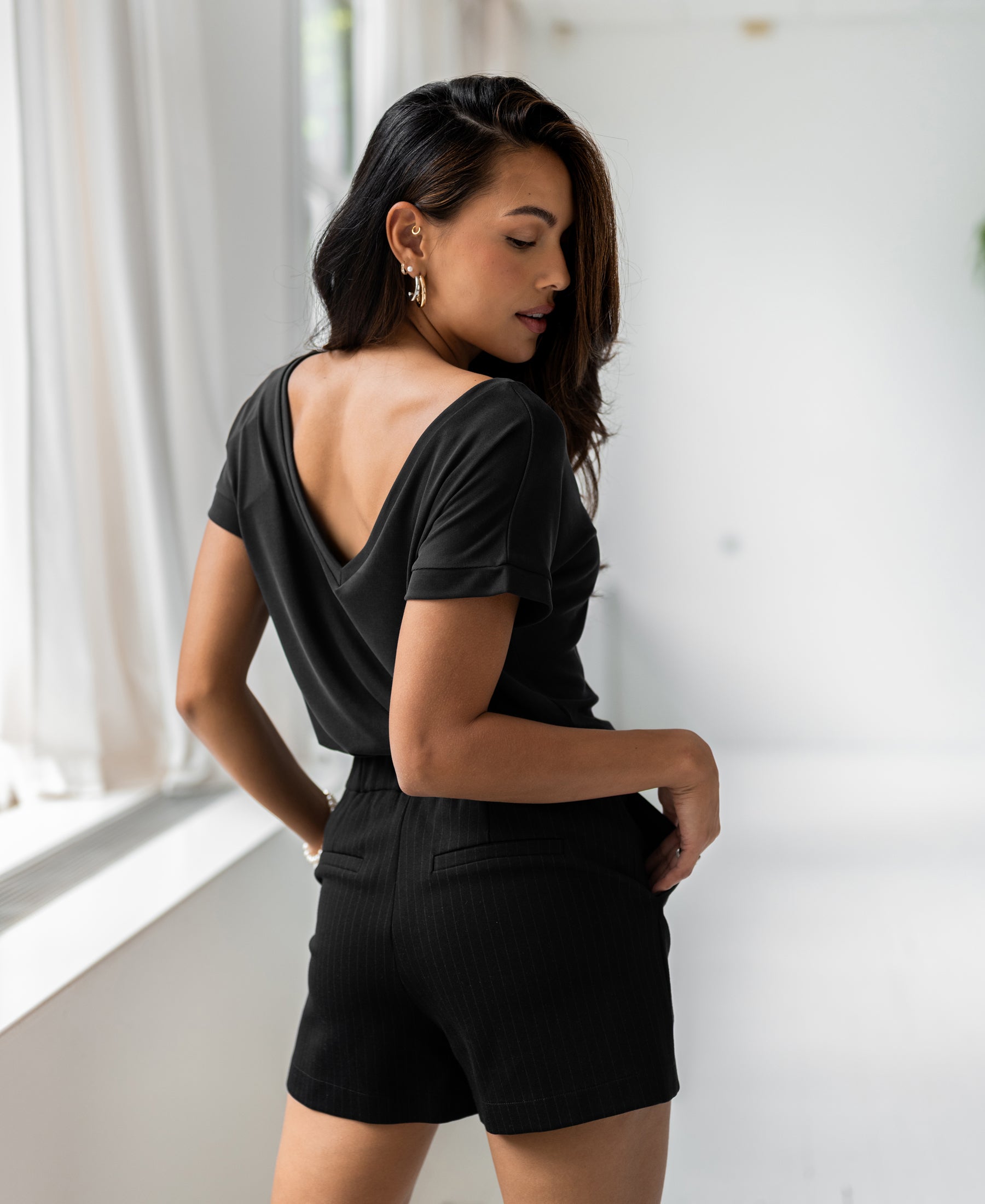 Woman wears PLEIN PUBLIQUE's LA JULIEN Charcoal two-way T-shirt, looking over her shoulder by a bright window.