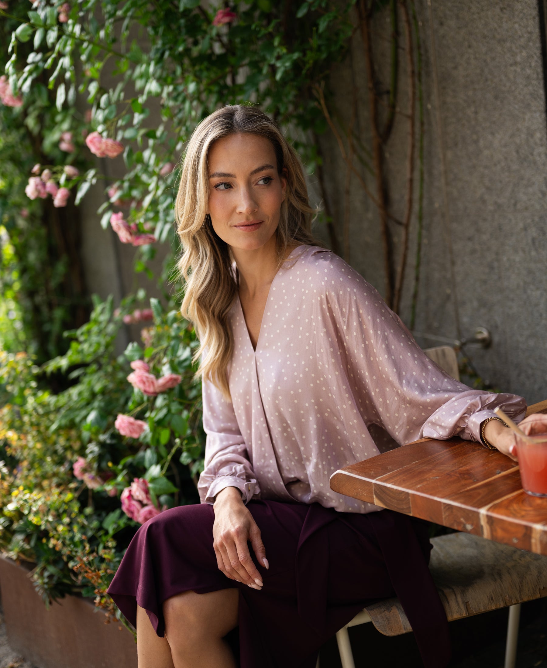 Wrap blouse with dot pattern in dusty pink and ivory, shown outdoors near pink flowers.