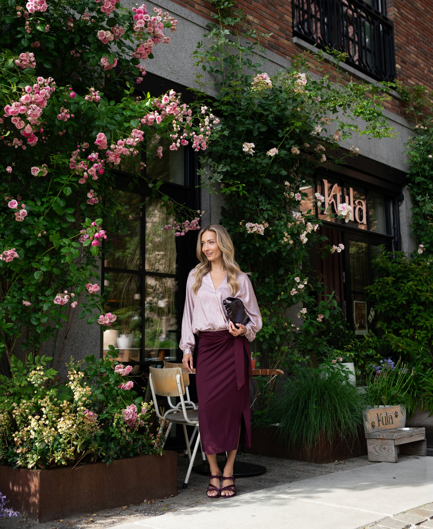 Woman in dusty pink polka dot wrap blouse stands outside a café with pink roses.