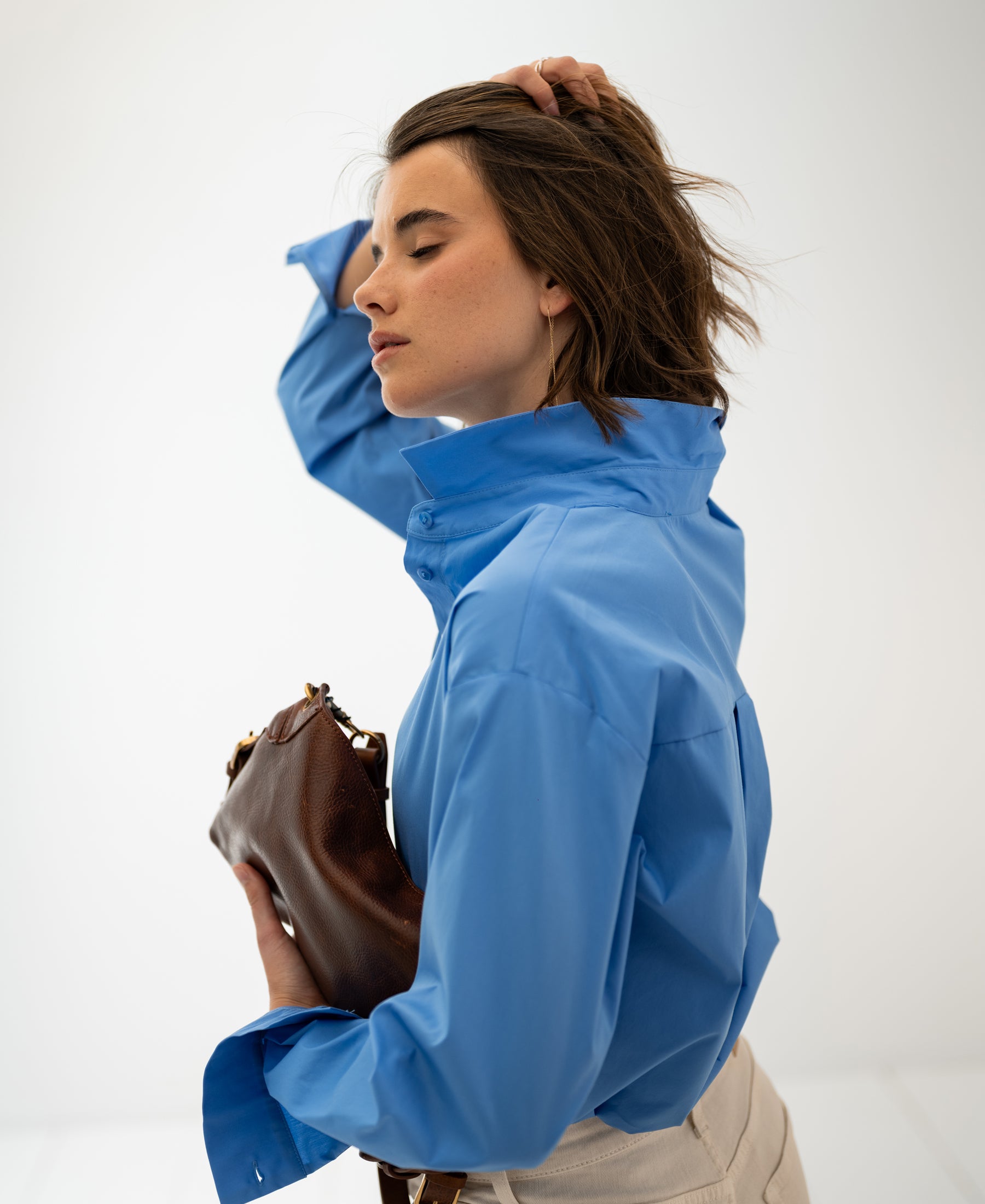 Woman wears oversized easy-iron blue blouse called Le Lille, posing with eyes closed against a white background.