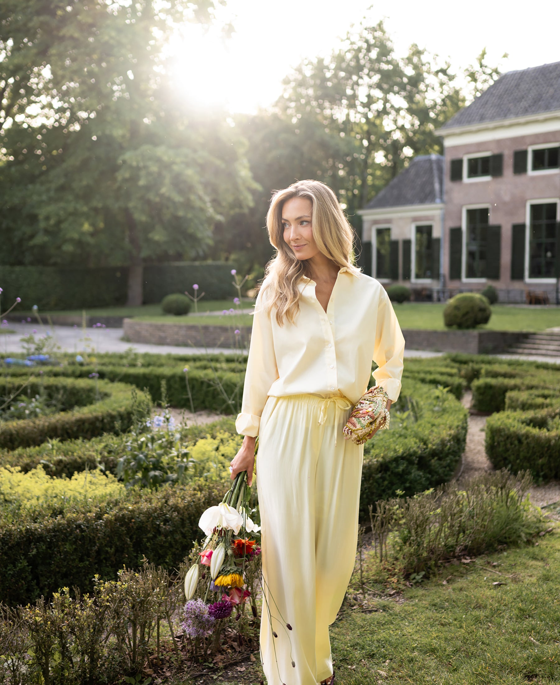 Light yellow LA LYNETTE trousers by PLEIN PUBLIQUE, shown in a sunlit garden with a house in the background.