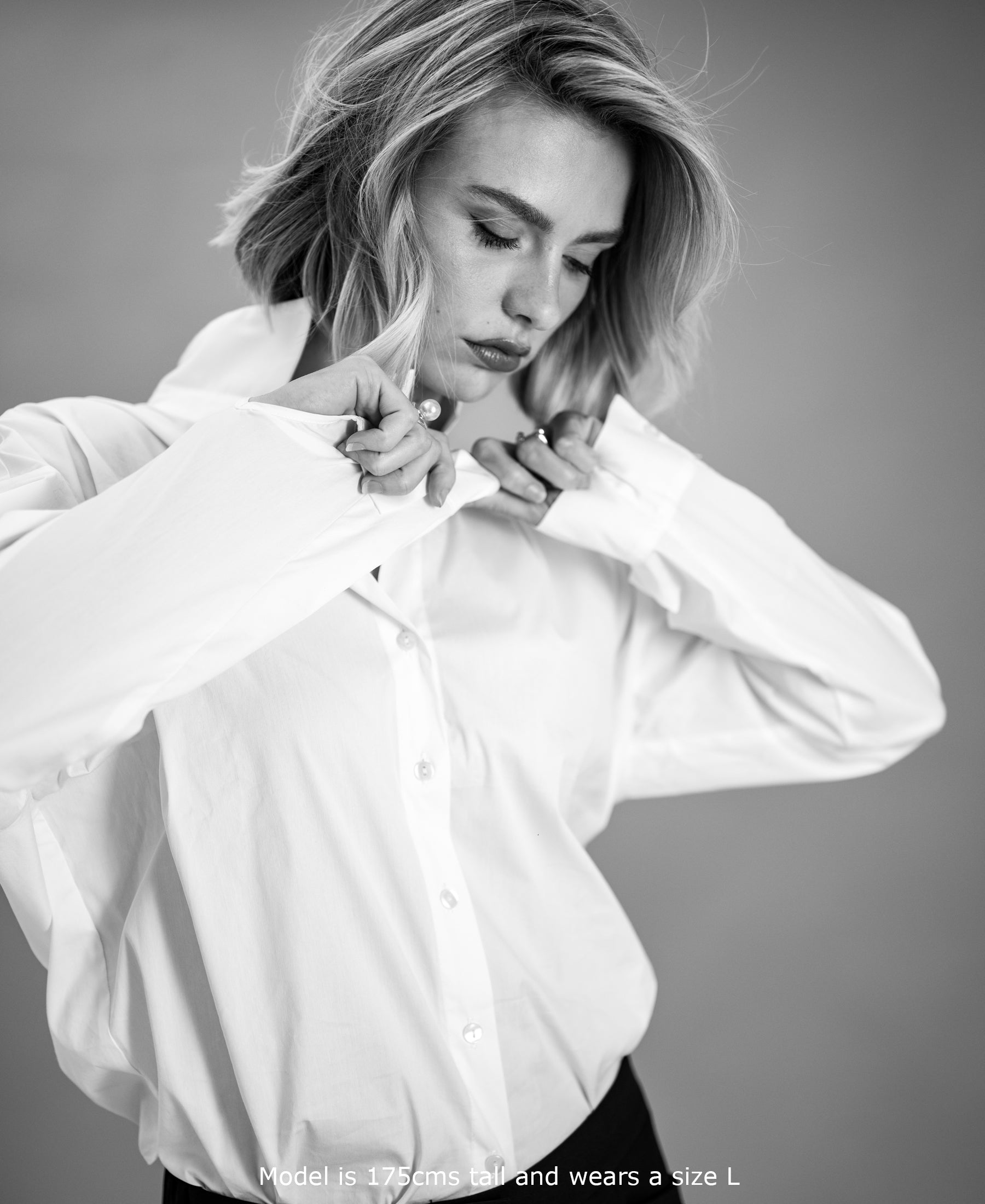 Black and white photo of the Plein Publique oversized white blouse, with model buttoning the front, seen from above.