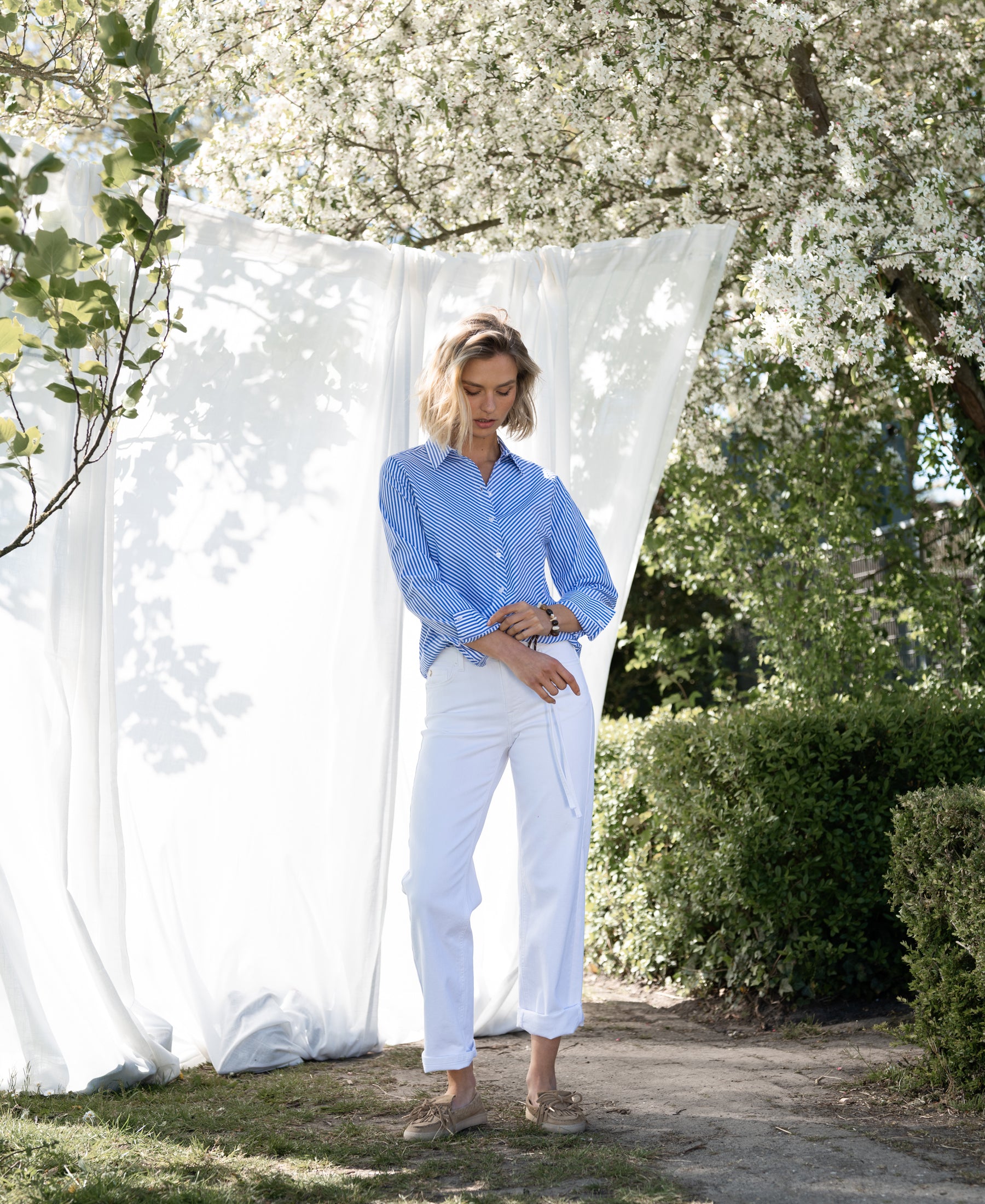 Striped white-blue blouse by PLEIN PUBLIQUE LA LOIRE shown outdoors with blooming trees in the background.