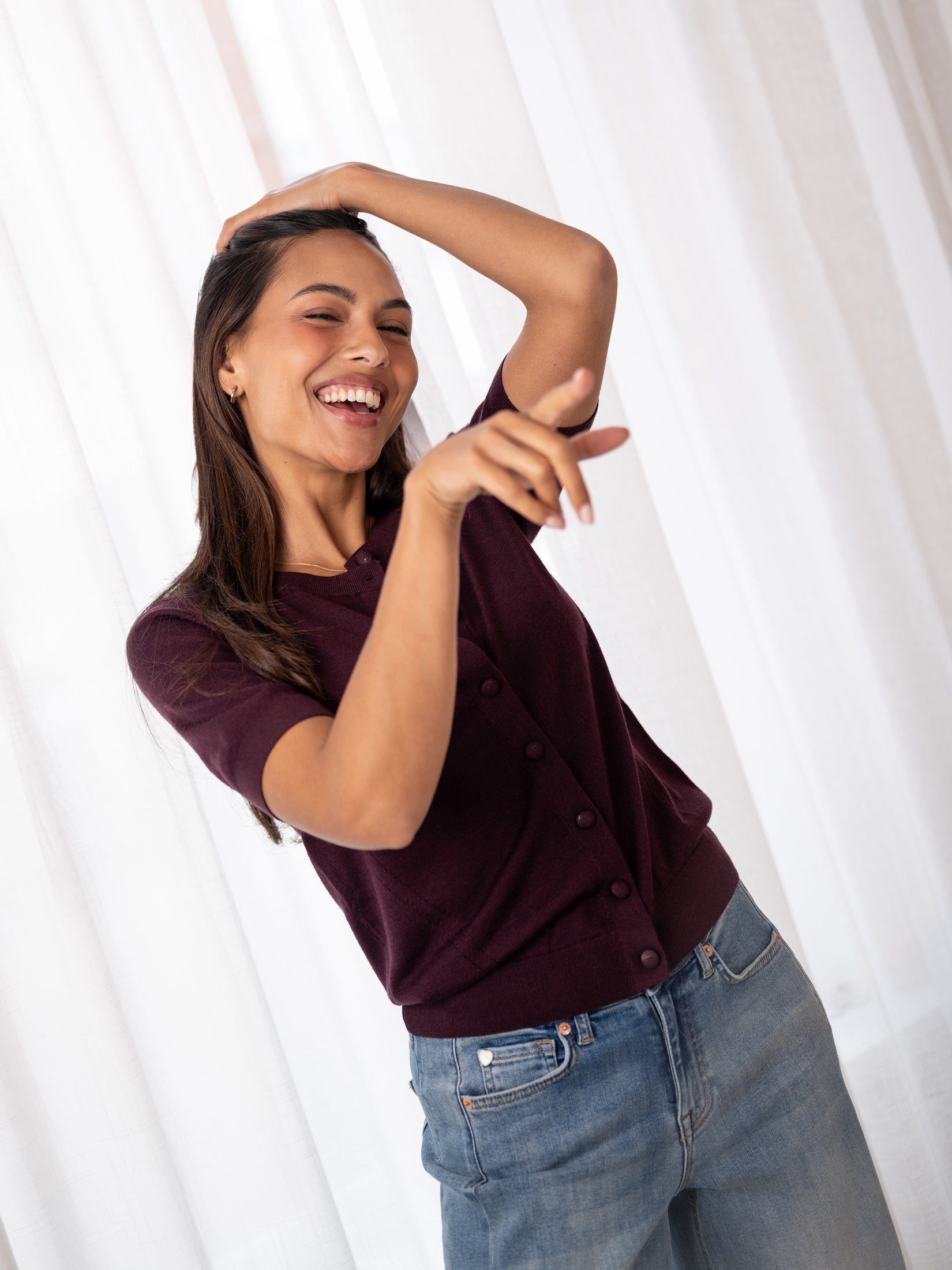 Woman wearing the PLEIN PUBLIQUE LA LORAINE plum merino wool silk cardigan raises one arm and smiles.
