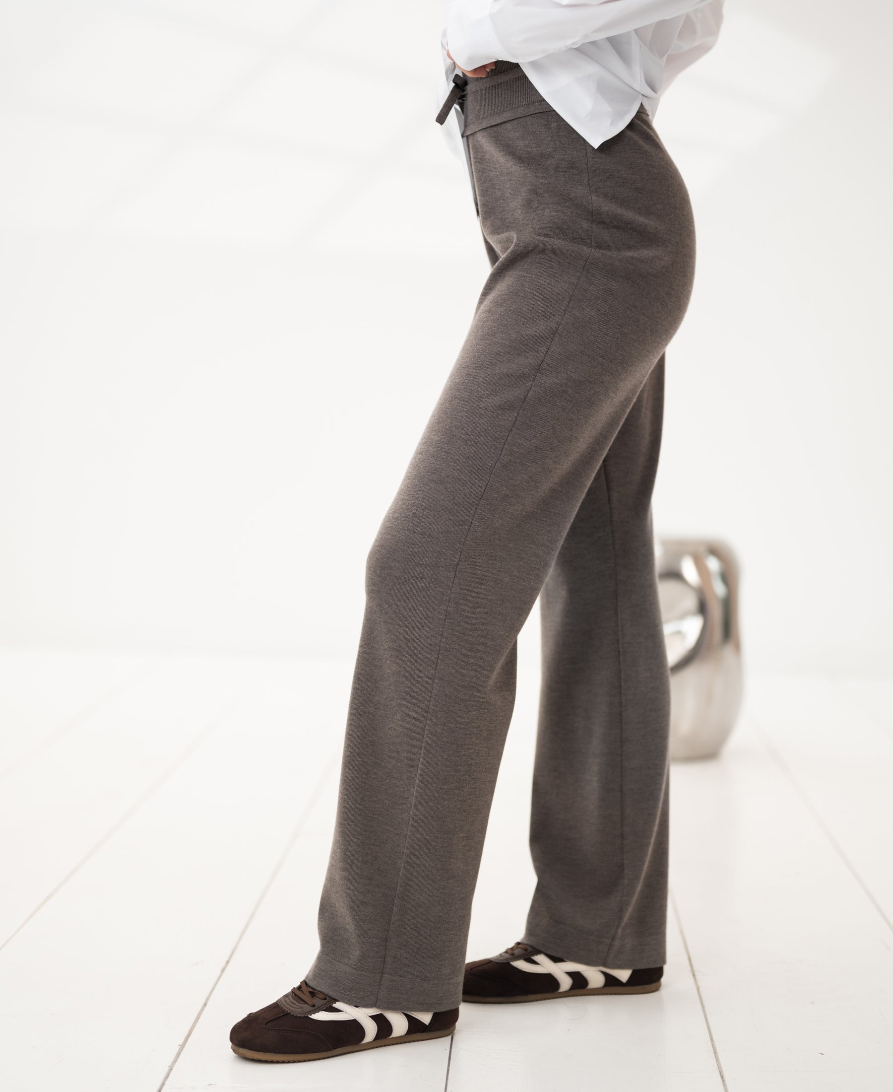 Woman wearing dark sand knit trousers stands by a modern stool.
